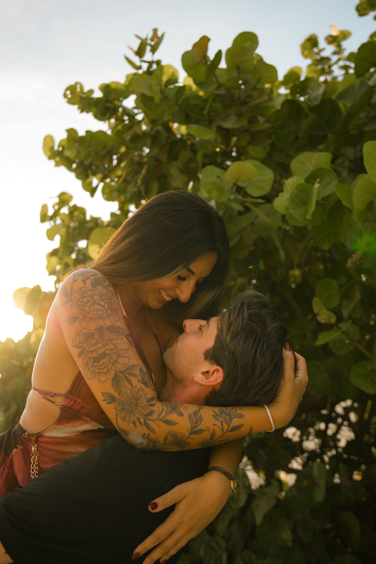 Smiling woman with floral arm tattoos embraces her partner in warm golden sunlight in front of green leafy foliage.