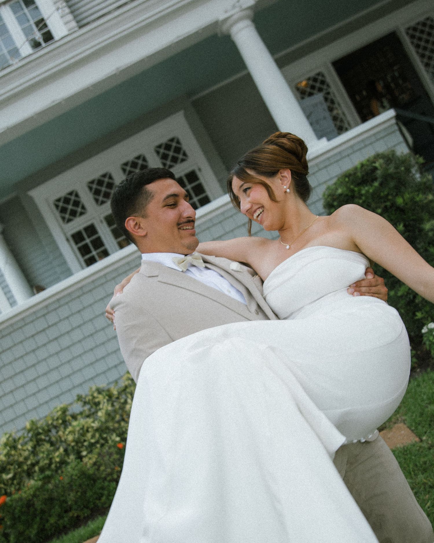 Groom in a tan suit smiles while carrying his laughing bride in a white strapless gown outside a historic home.