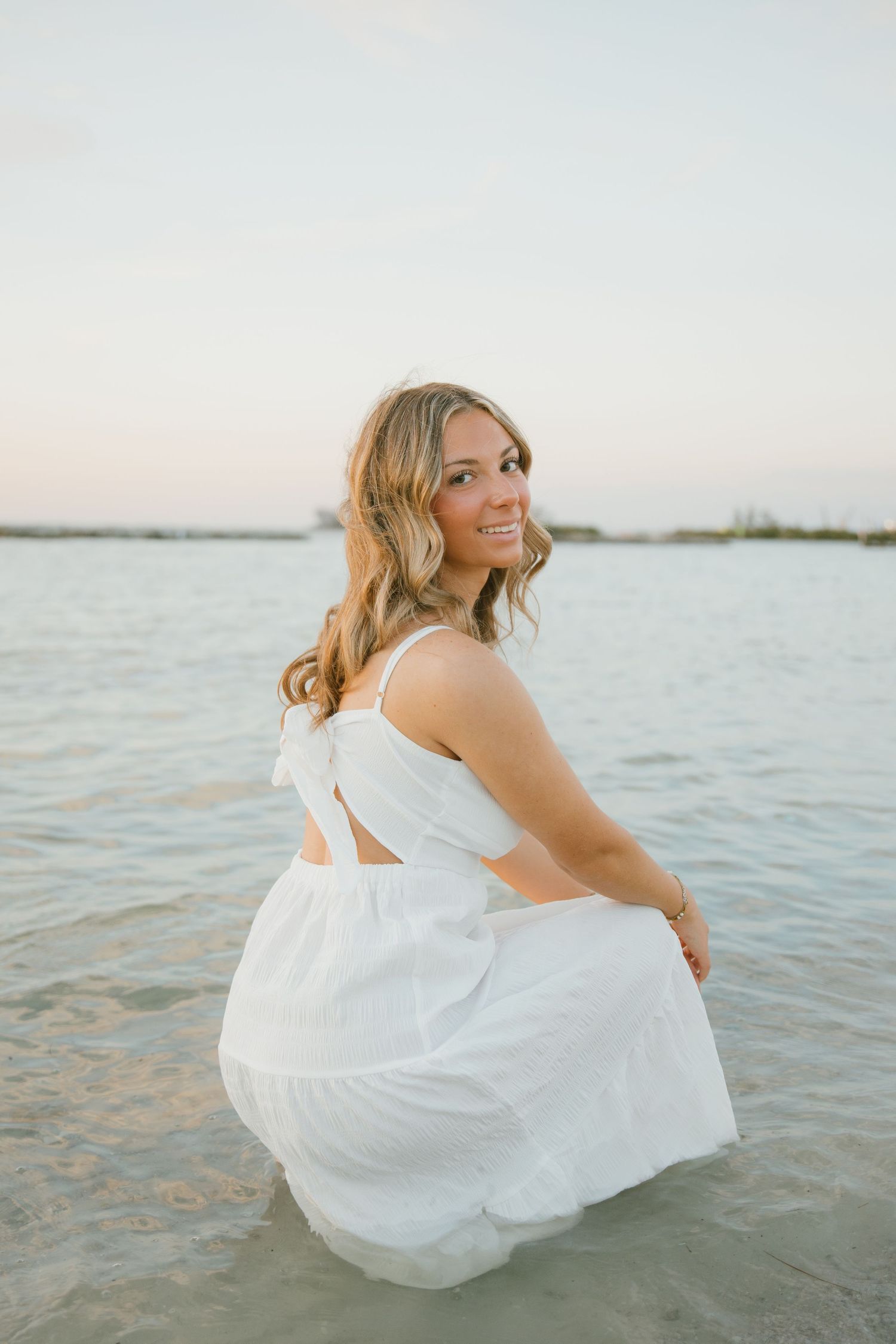 Smiling woman in a white tiered dress crouches in shallow water and looks back over her shoulder at dusk.