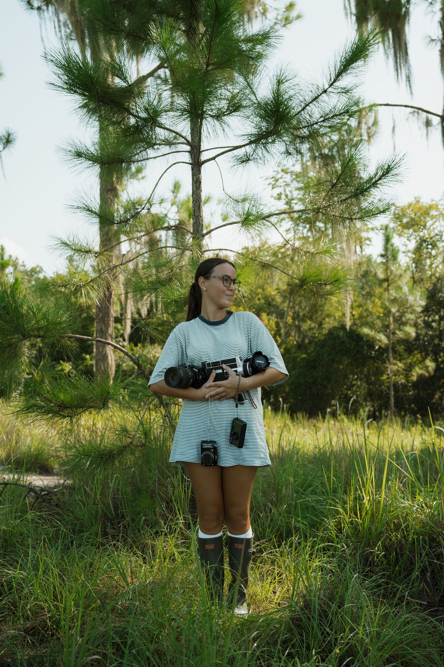 Woman in a light blue t-shirt dress and rubber boots holds several cameras while standing in tall grass near pine trees.