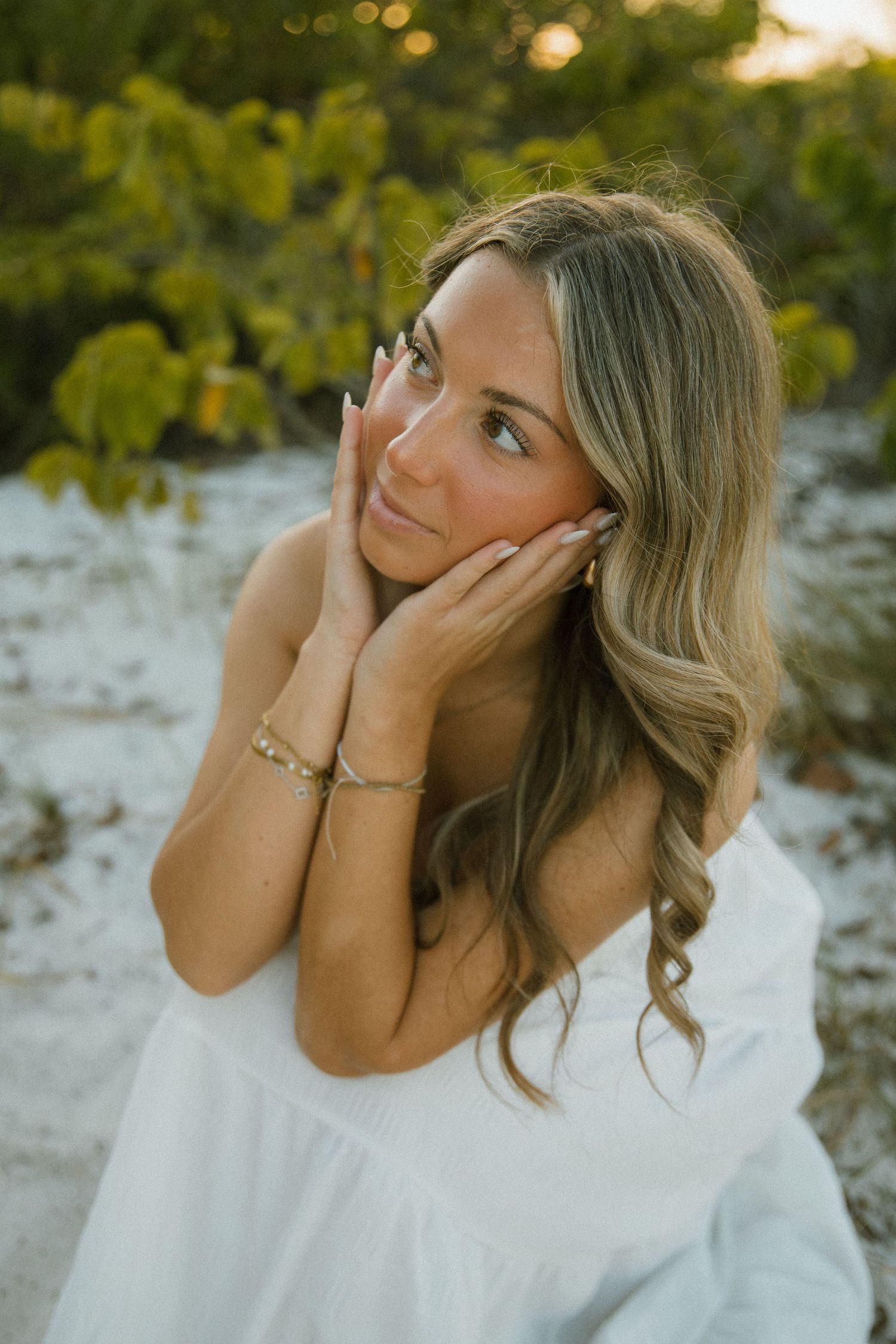Close-up portrait of a woman with wavy hair in a white dress, resting her face on her hands on the beach.