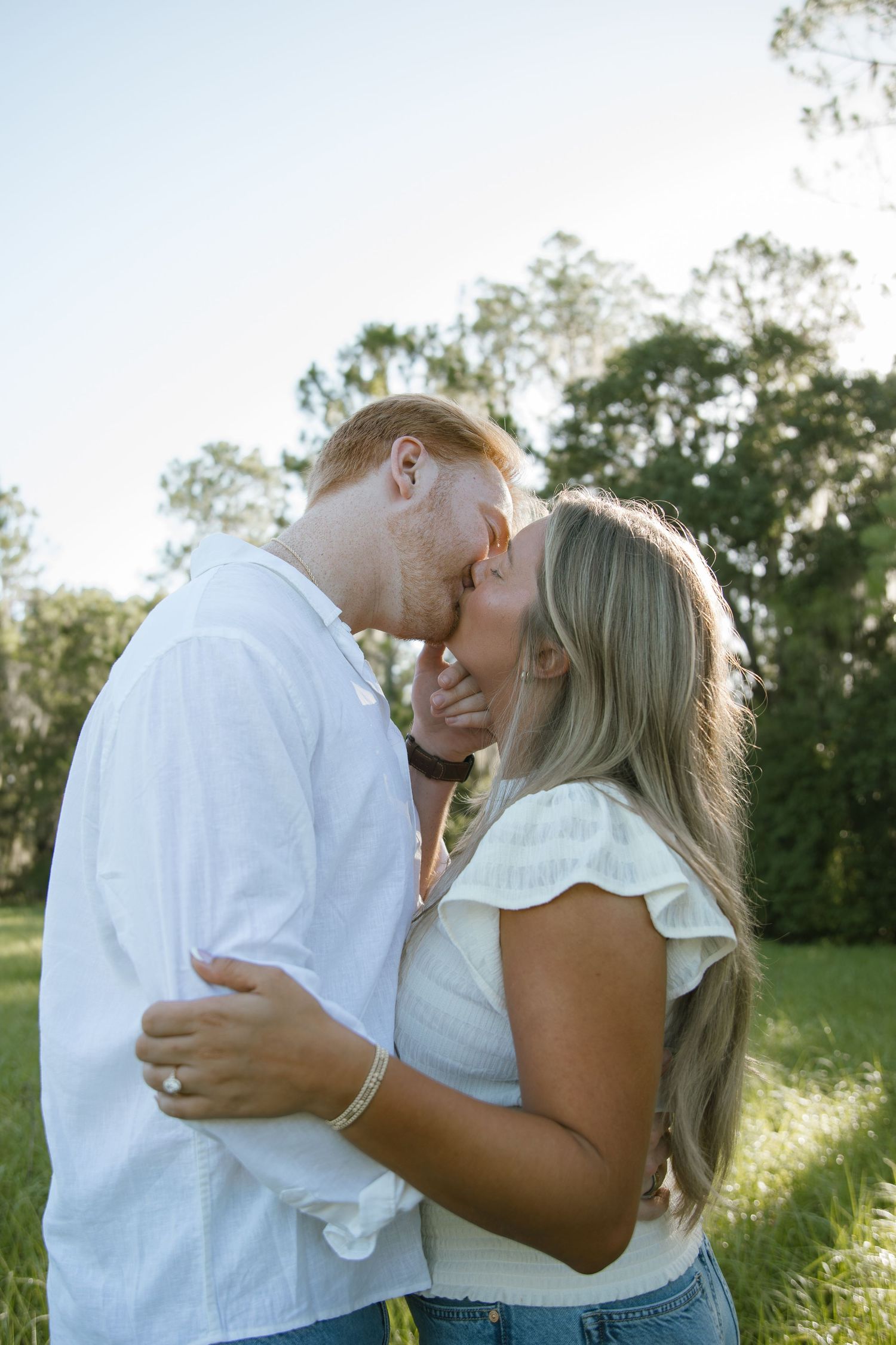 Engaged couple in white shirts share a kiss in a sunny grassy field with trees in the background.