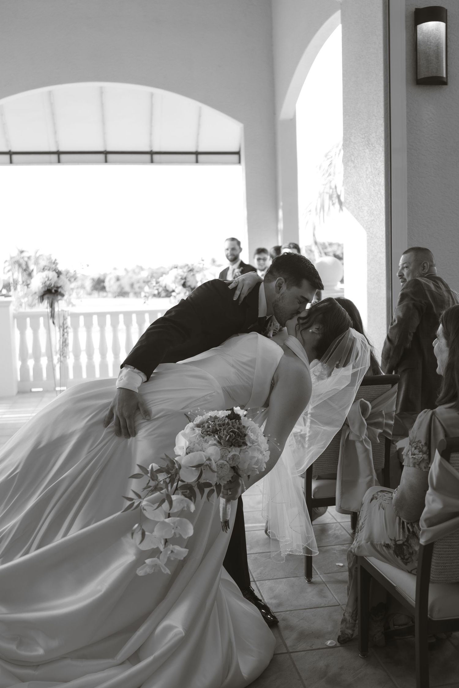 Groom dips and kisses his bride in a black and white photo on an arched terrace as wedding guests watch.