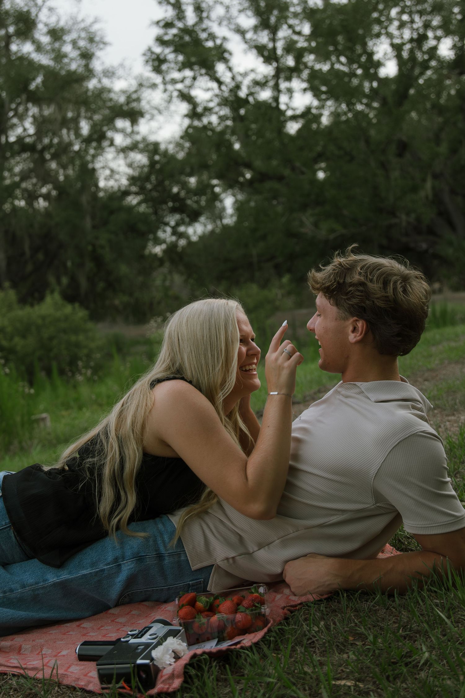 Couple laughs on a picnic blanket in a green meadow, with strawberries and a vintage camera beside them.