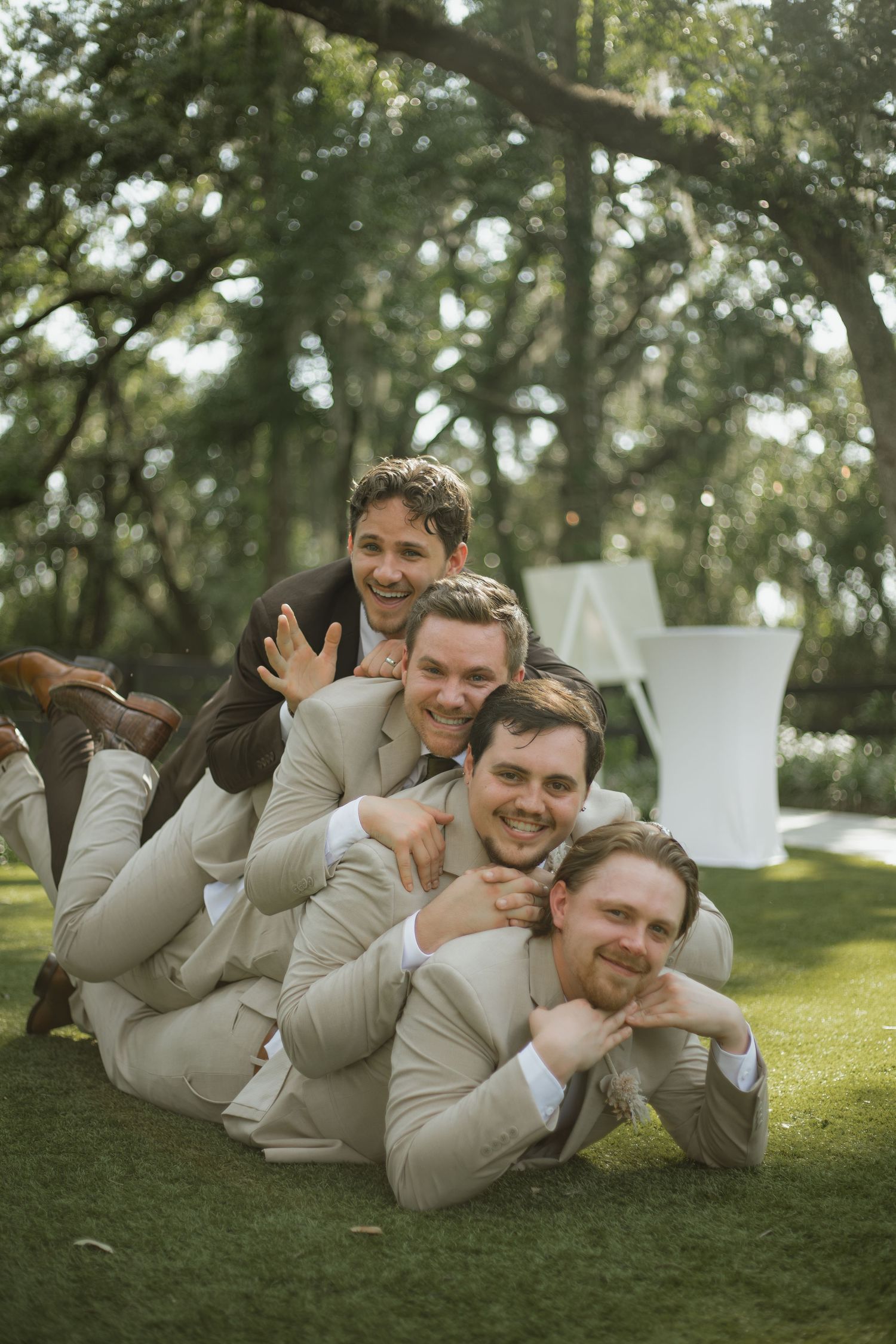 Four smiling groomsmen in tan suits lie stacked playfully on the grass, with the groom in a brown suit on top at a wedding.