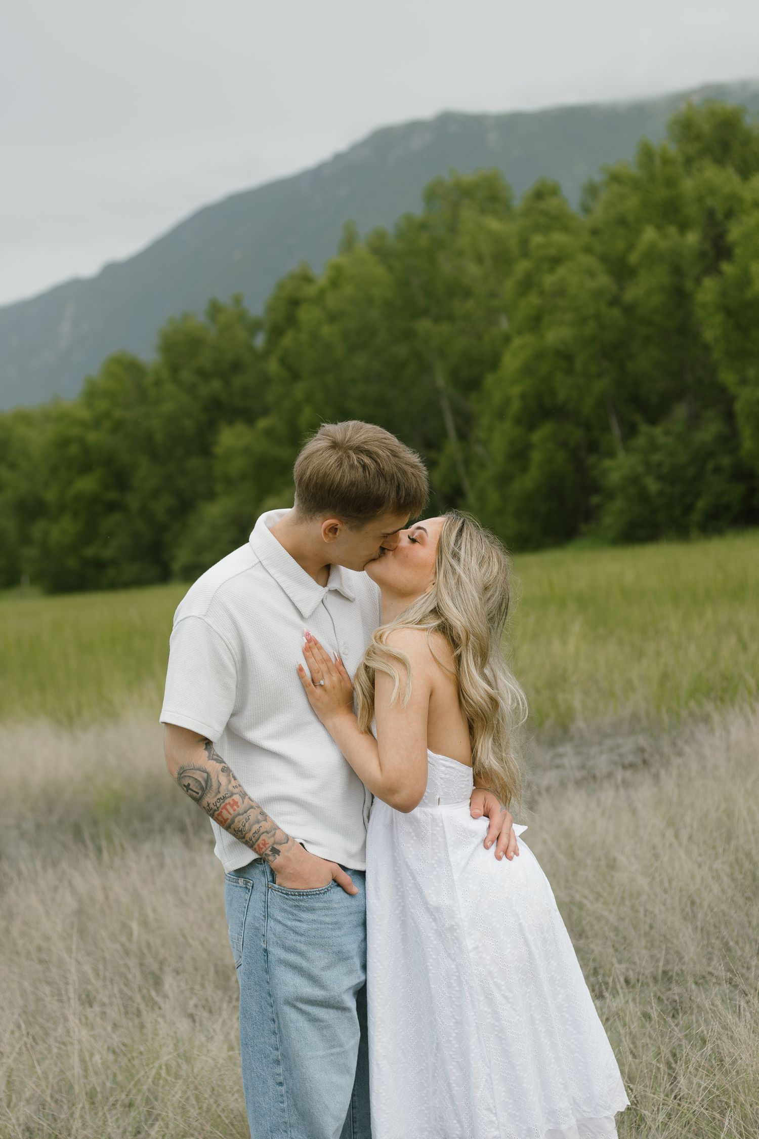 Couple kissing in a grassy meadow with green trees and mountains behind, she wears a white dress and he has an arm tattoo.