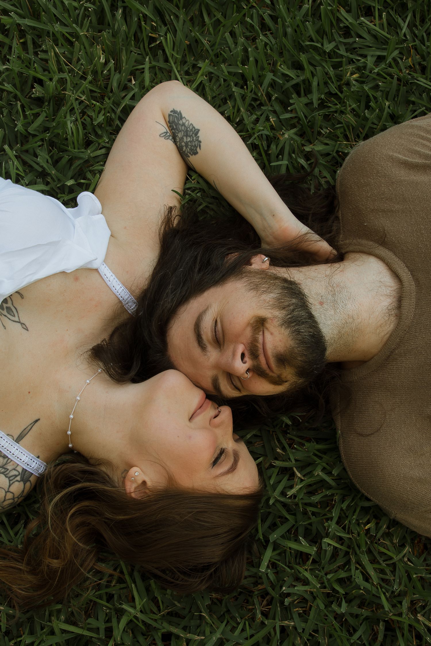 Couple lying head to head on green grass, smiling with eyes closed in a romantic outdoor portrait.
