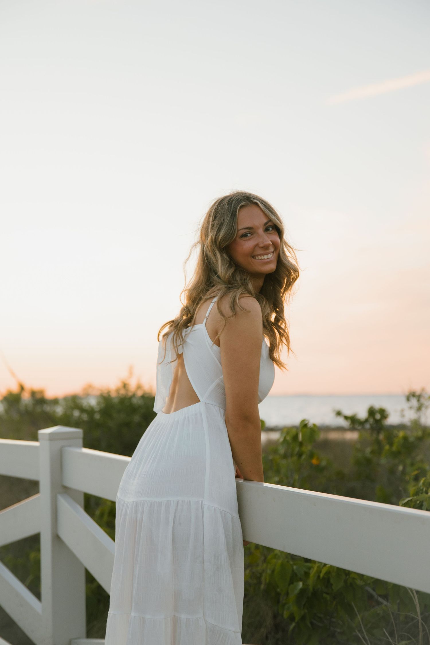 Smiling woman in a white open-back dress leans on a white fence with pastel sunset skies and sea behind her.