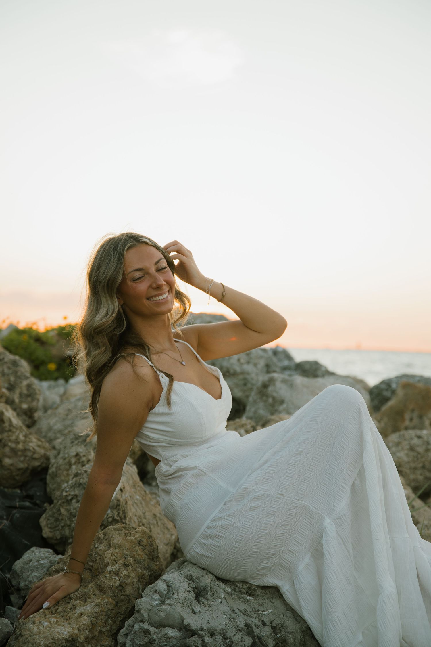 Smiling woman in a white dress sits on rocks by the water, touching her hair as the sky glows at dusk.