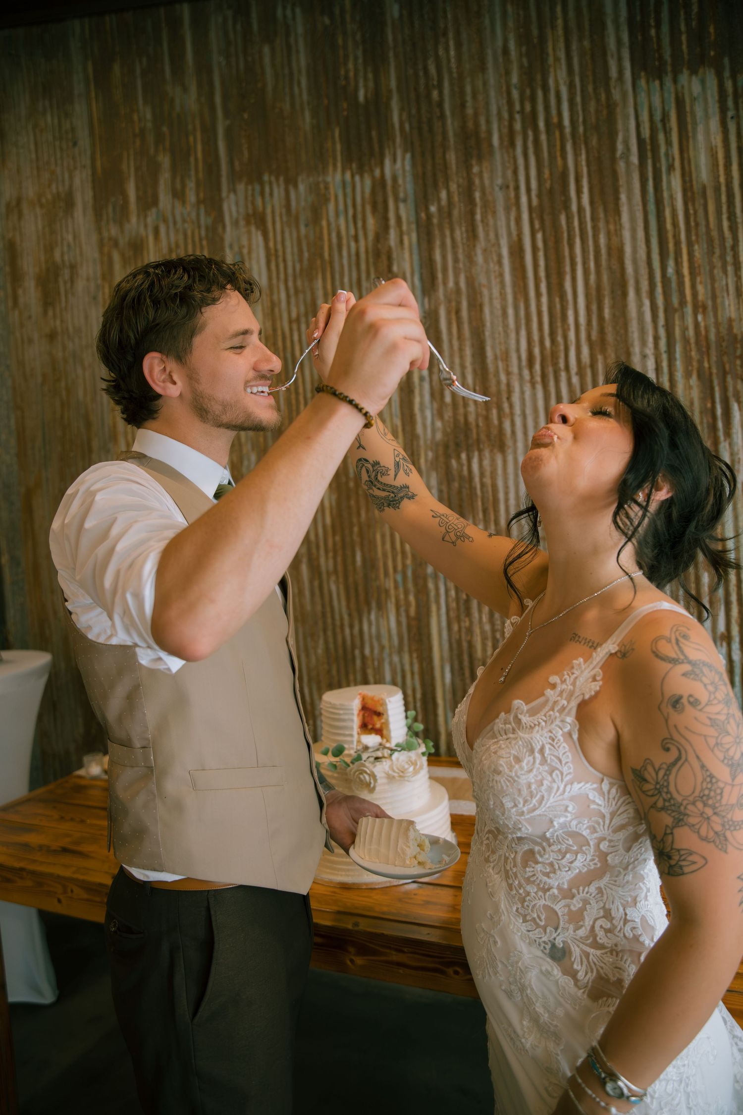 Smiling groom feeds wedding cake with a fork to his tattooed bride in a lace gown at a rustic reception.