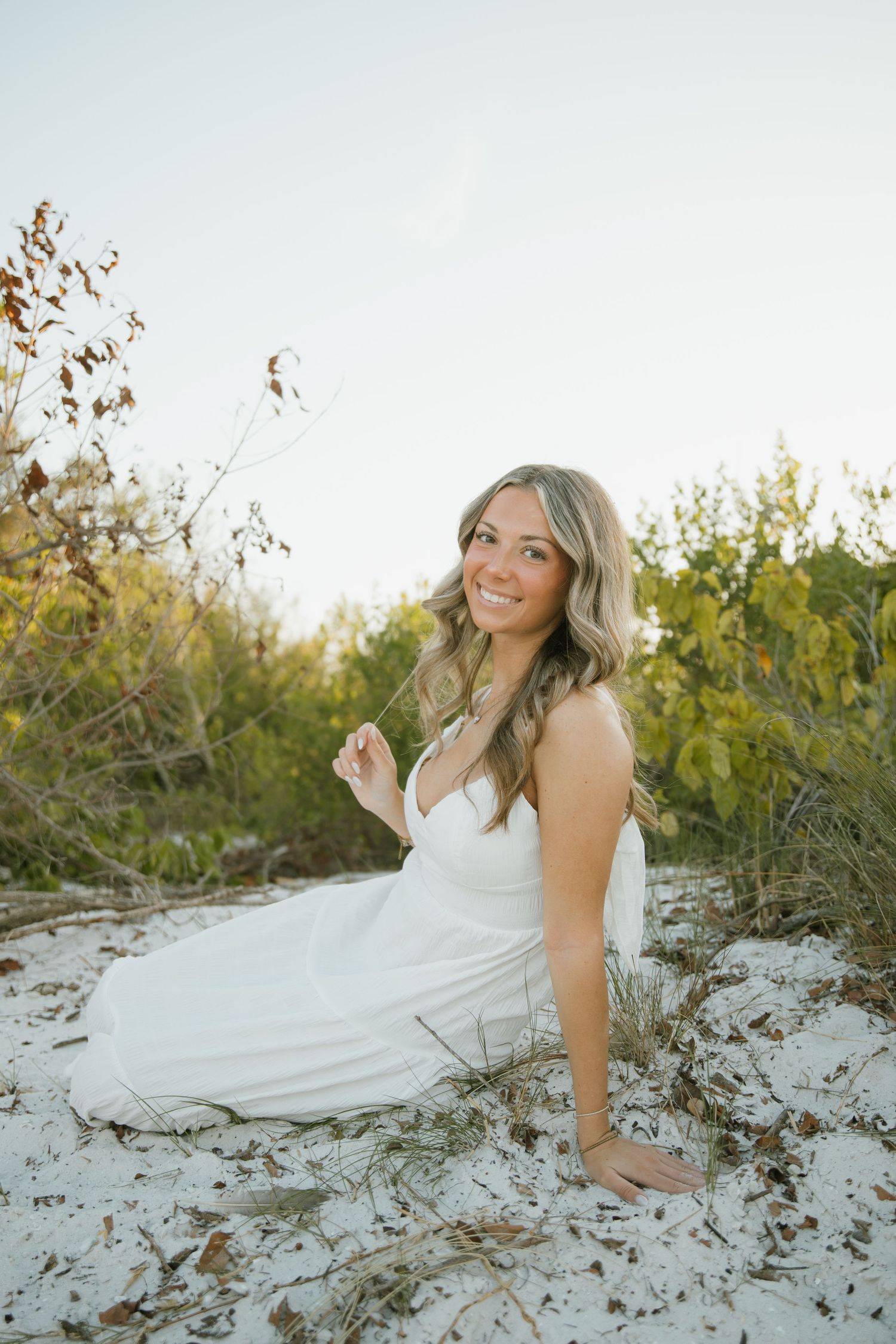 Smiling woman in a white dress sits on white beach sand with coastal shrubs and warm sunset light behind her.