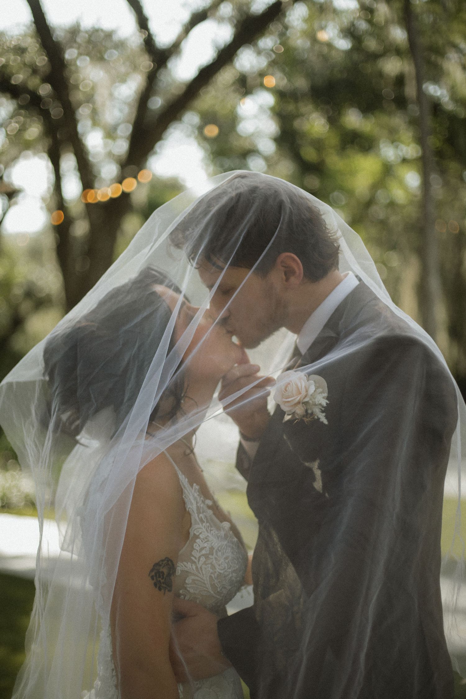 Bride and groom share a romantic kiss under a sheer wedding veil outdoors, with string lights glowing in the trees behind them.