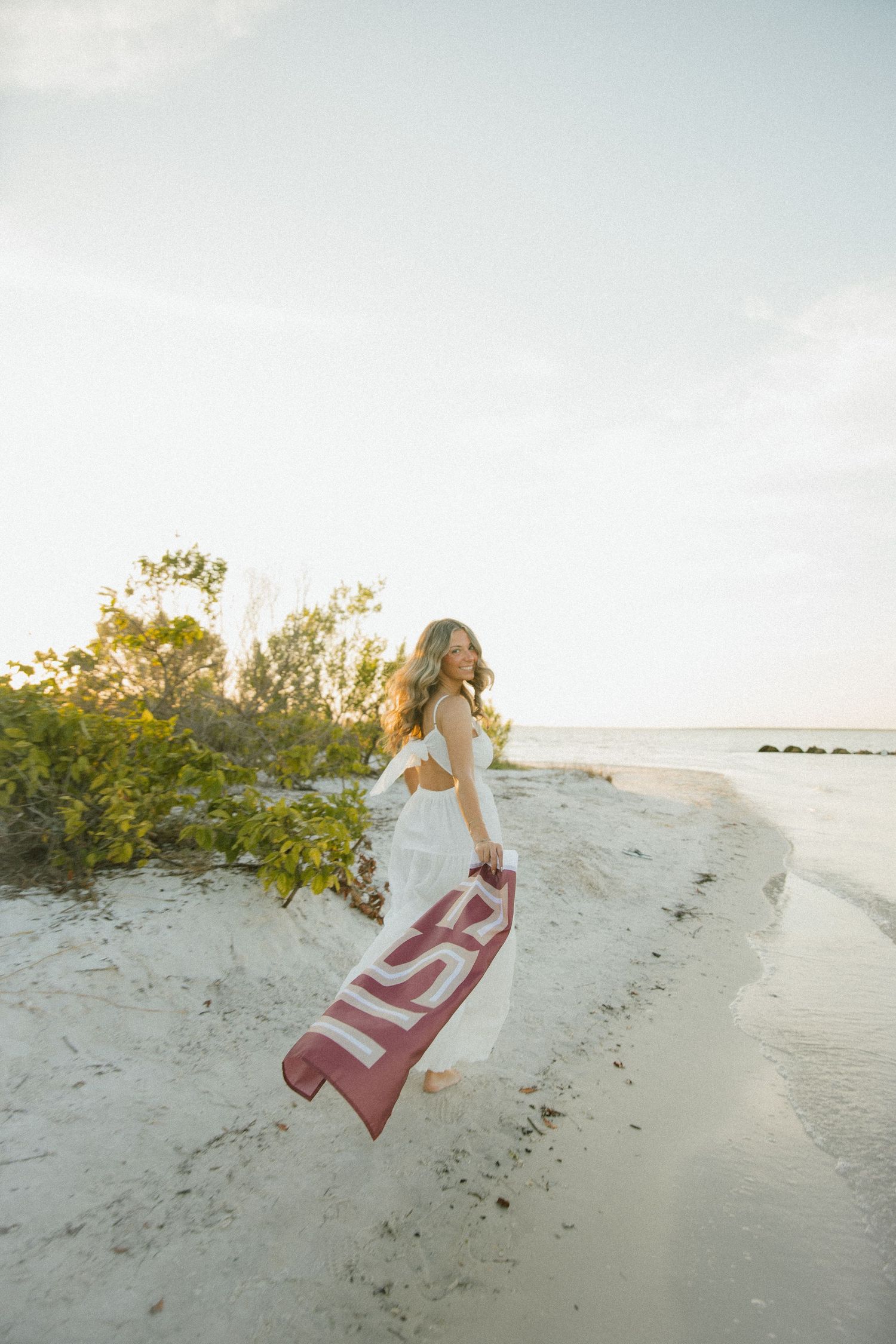 Woman in a white dress glances back over her shoulder, trailing an FSU flag across a sunlit sandy beach.