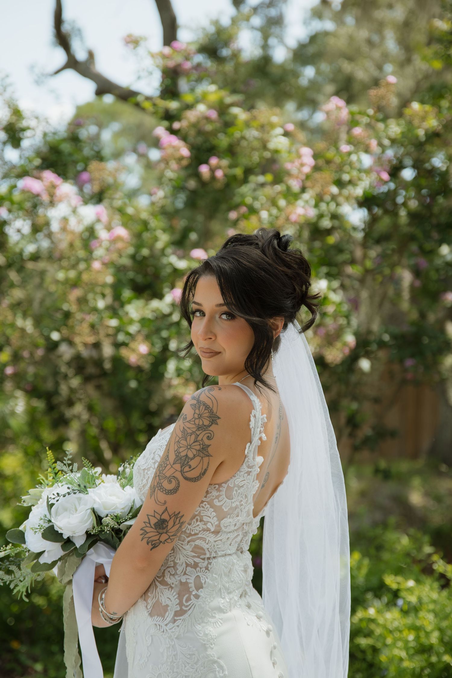 Bride in a lace wedding gown and long veil holds a white floral bouquet, showing her arm tattoo while posing by blooming pink garden roses.