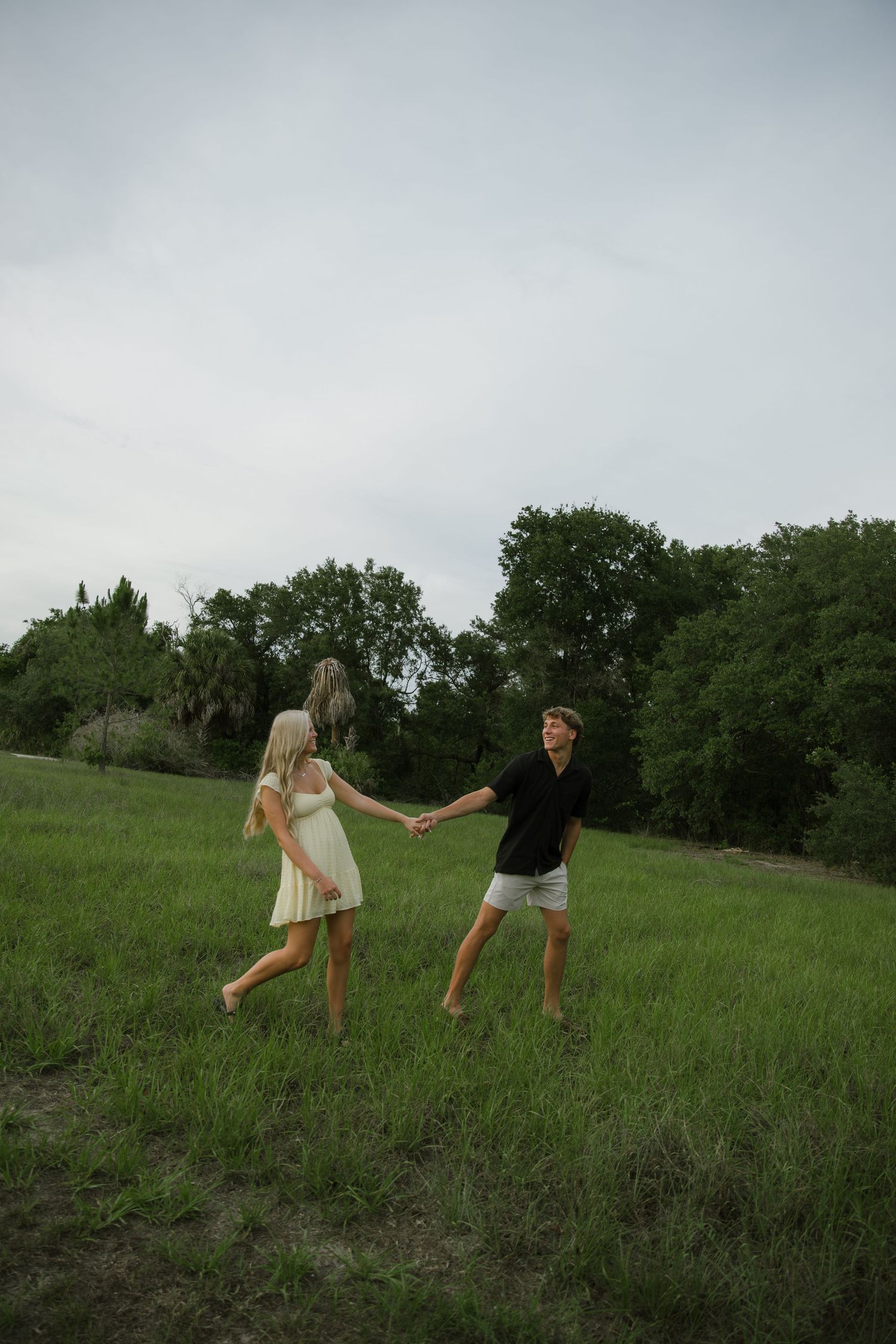 Barefoot couple holding hands and twirling in a green grassy field with trees under a cloudy sky.