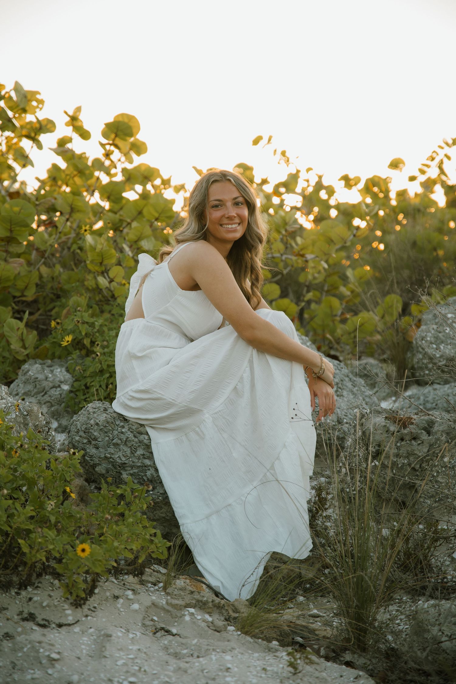 Smiling woman in a white sundress crouches on coastal rocks beside green sea grape bushes at golden hour.