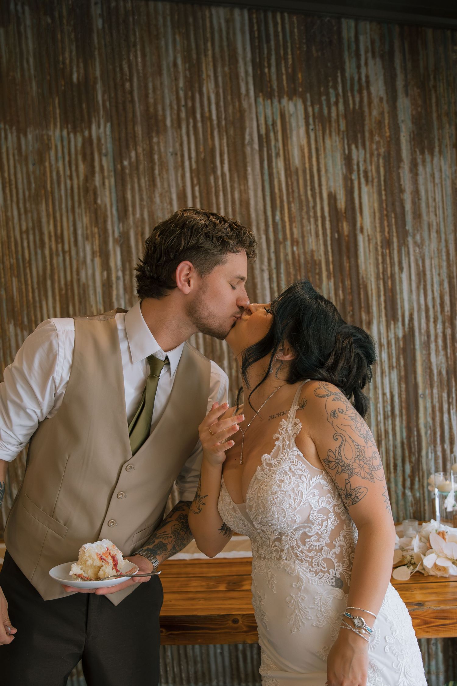 Bride and groom share a kiss while holding a slice of wedding cake against a rustic corrugated metal wall.