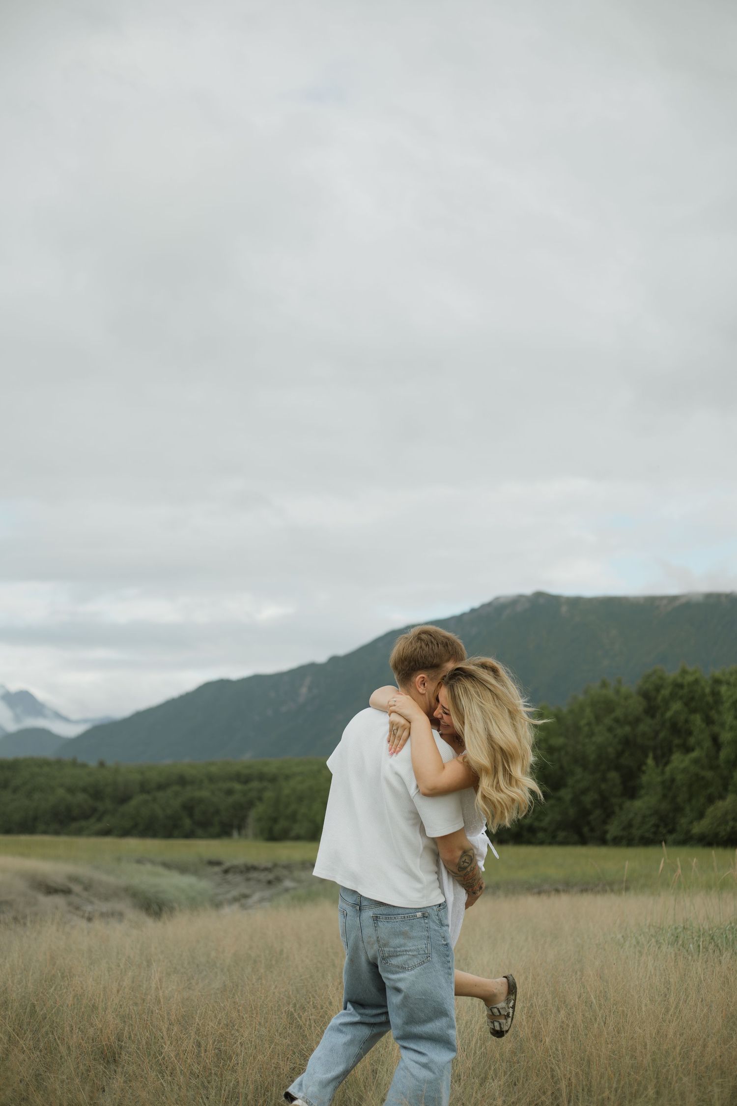 Couple embracing in a golden grass field with green mountains and a cloudy sky behind them, the woman lifted in a joyful hug.
