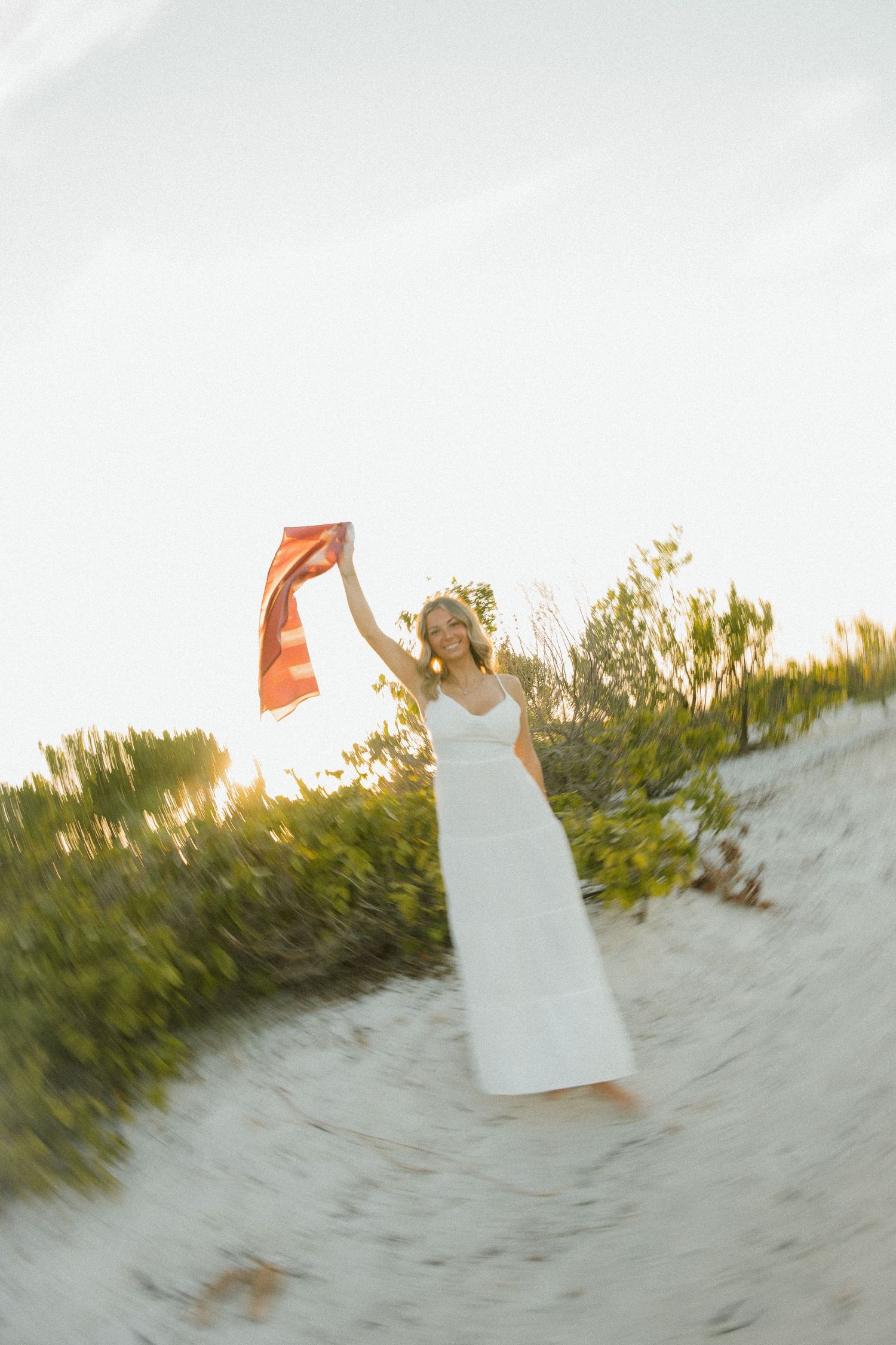 A woman in a white dress holds an orange cloth high in the air while standing on a sandy dune at sunset.