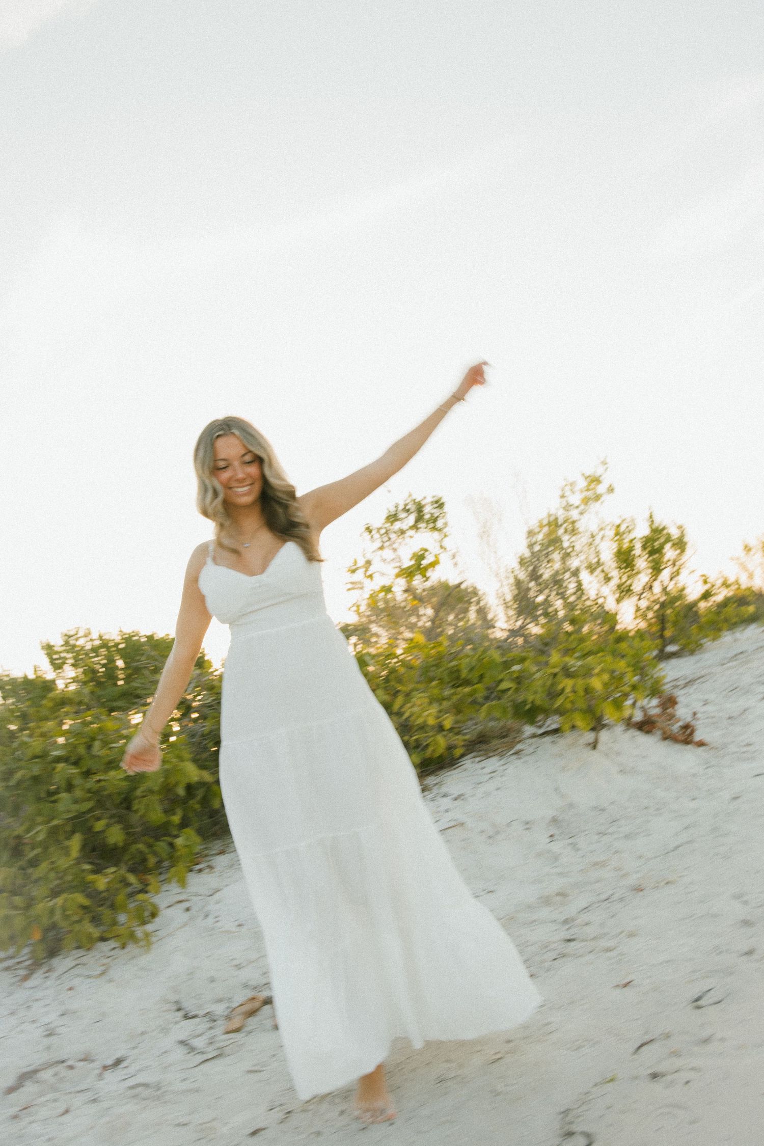 Woman in a flowing white dress dances with one arm raised on a sandy dune beside green coastal shrubs.