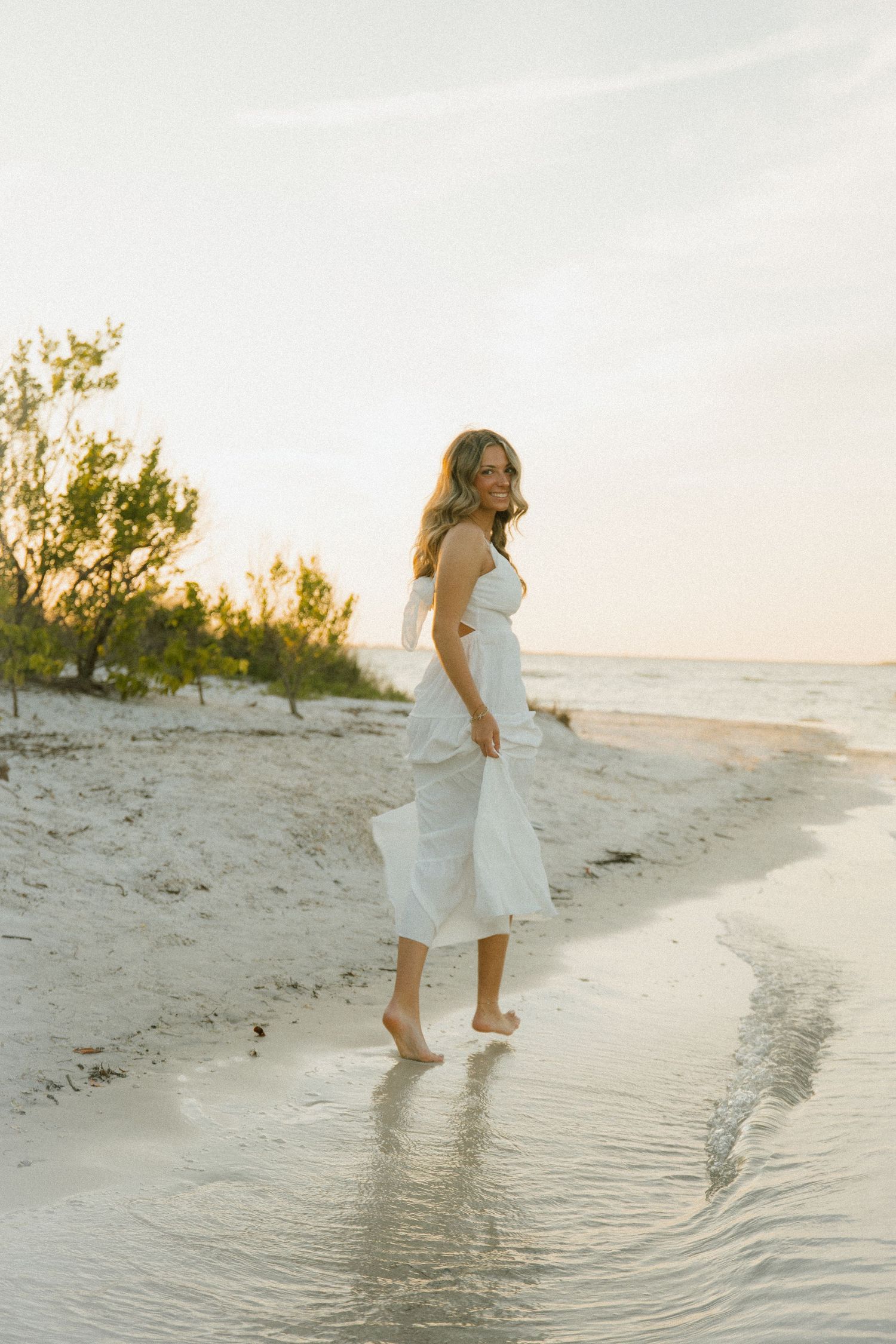 Barefoot woman in a white maxi dress walks along the shallow water's edge of a sandy beach at sunset.
