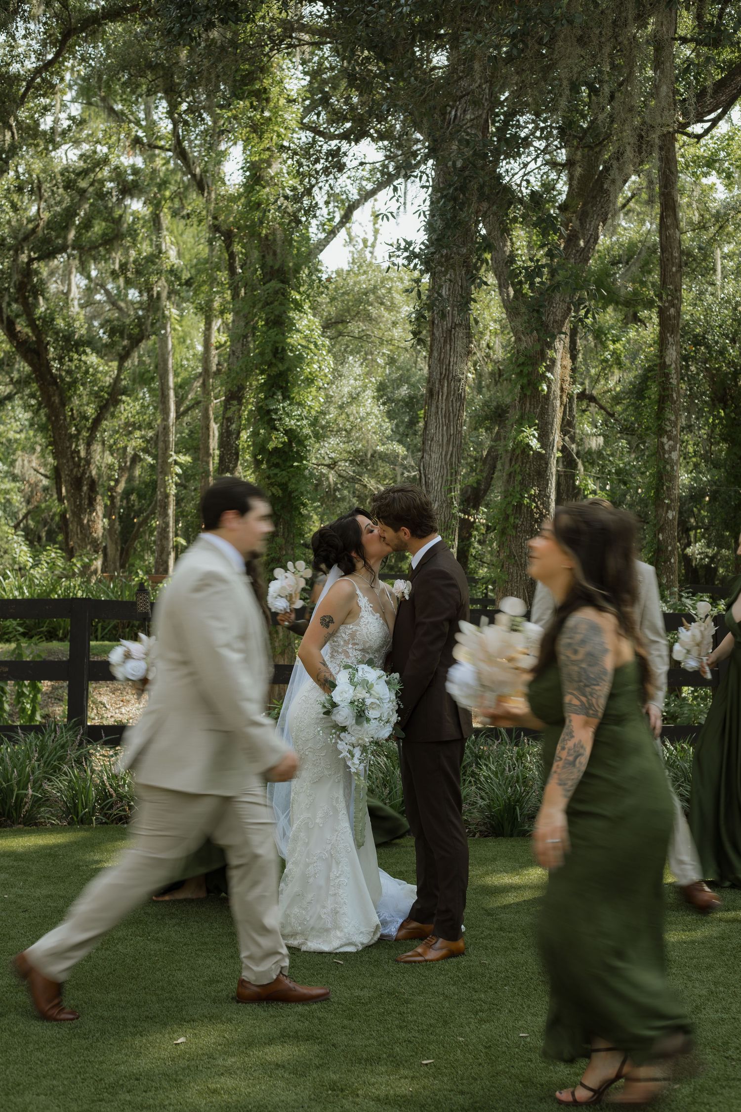 Bride and groom share a kiss on a lawn beneath moss-draped oak trees as wedding guests in motion walk past carrying bouquets.