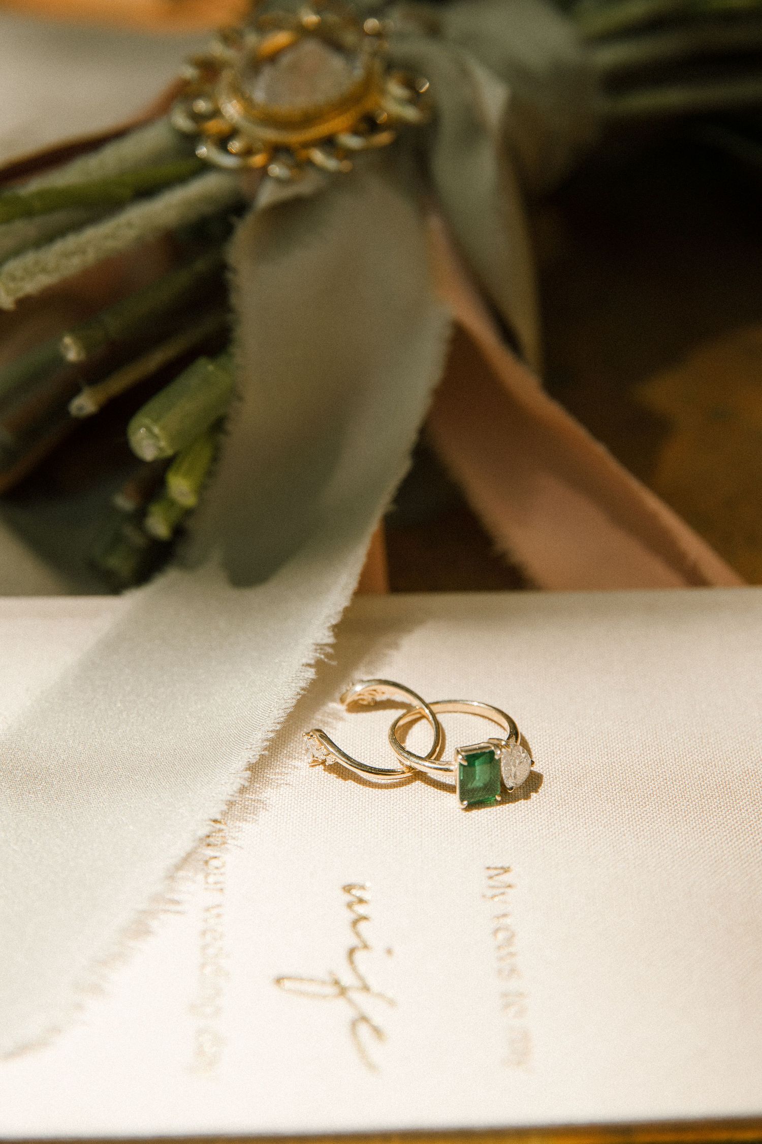 Emerald and diamond wedding rings rest on a cream invitation card beside a bouquet tied with a silk ribbon.