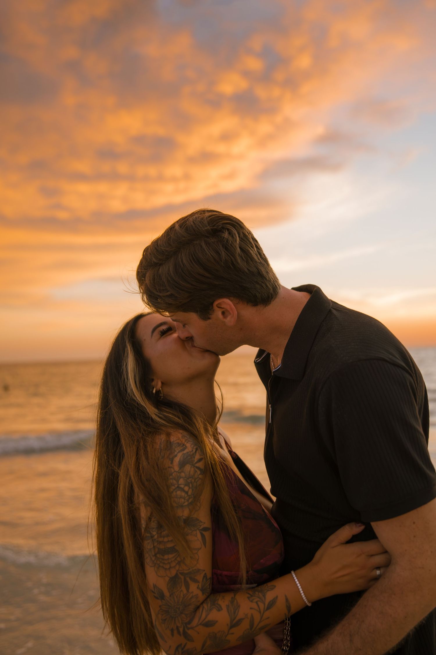Couple kisses on the beach at sunset under a vivid orange sky, the woman's tattooed arm wrapped around her partner.