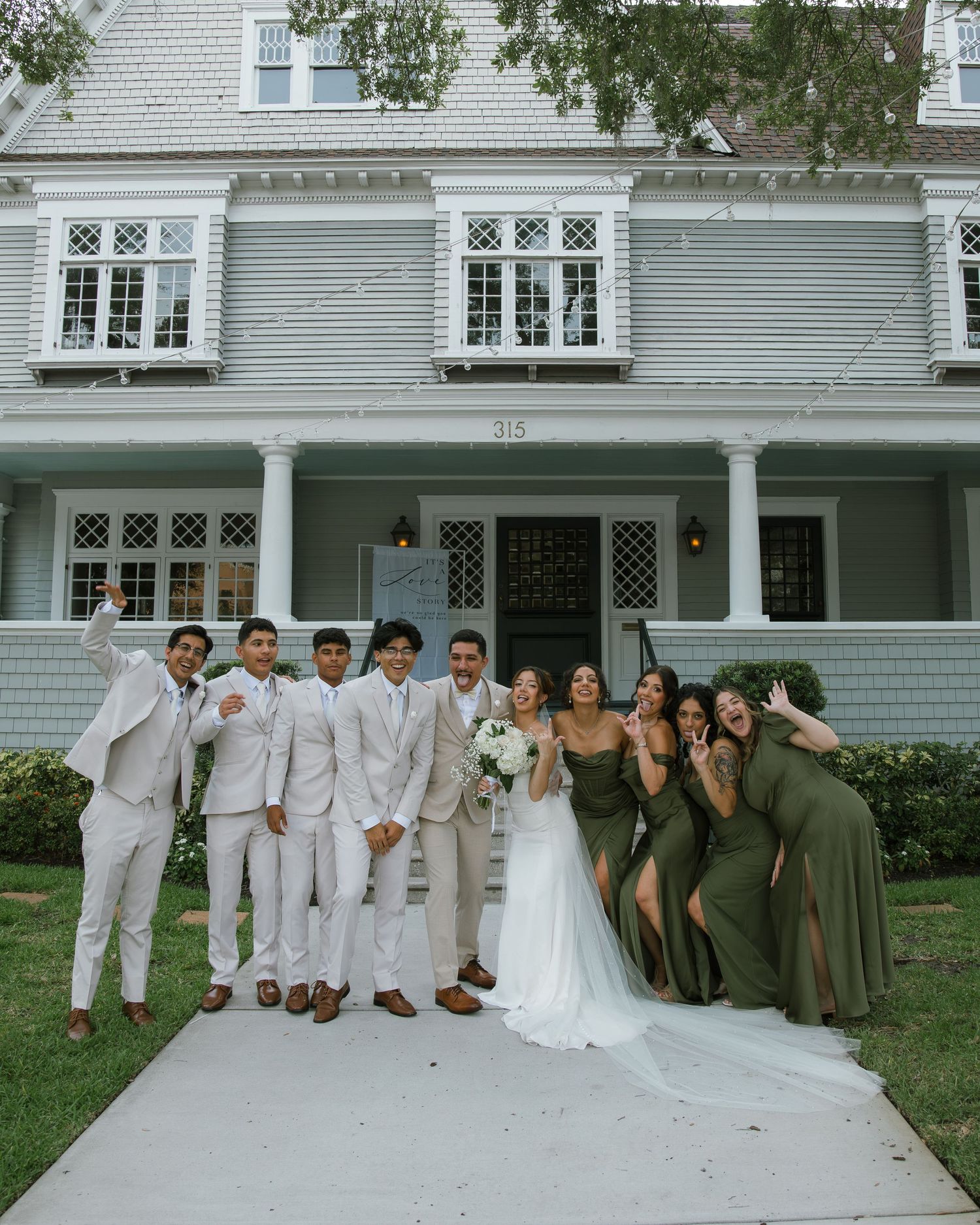 Wedding party of groomsmen in light suits and bridesmaids in olive green dresses cheer around a bride and groom outside a house.