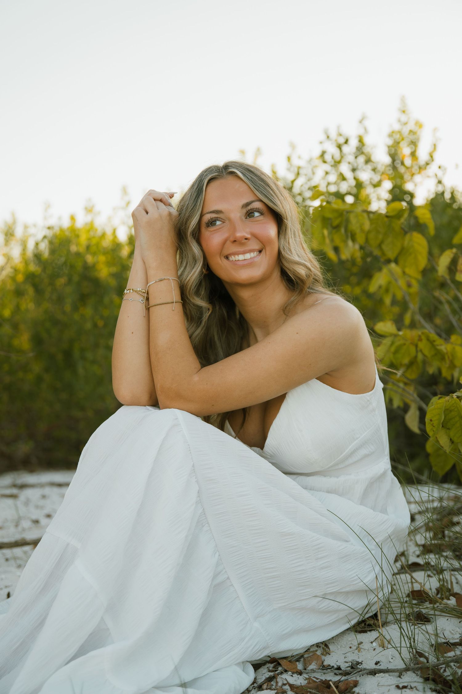A woman in a white sundress smiles warmly while sitting on pale sand with lush green coastal bushes in the background.