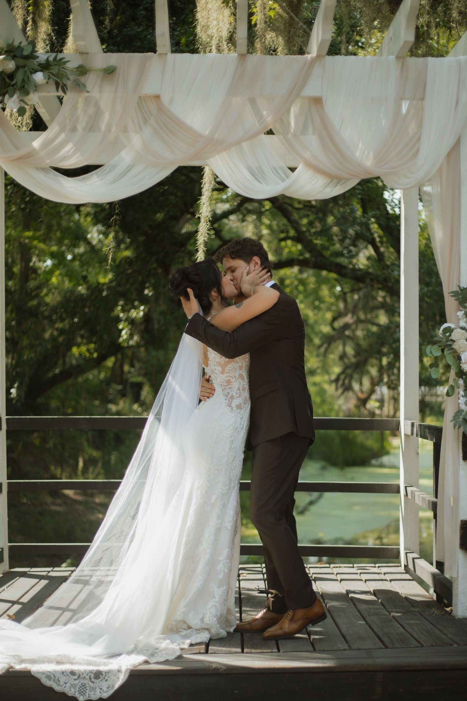Bride in a lace gown with a long veil kisses her groom in a brown suit beneath draped fabric on an outdoor wooden wedding deck.