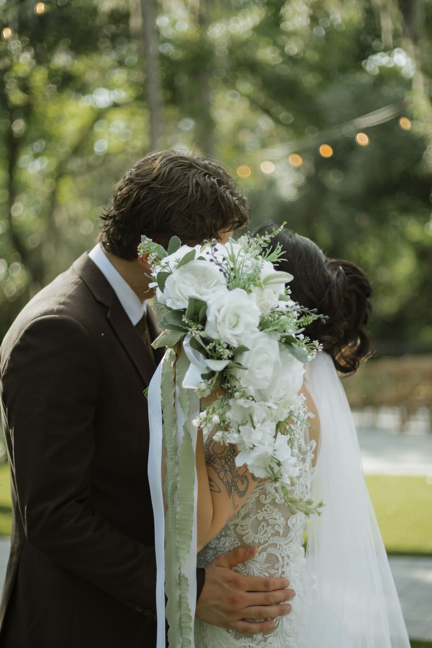 Groom in a brown suit kisses the bride behind a white and green bouquet with trailing ribbons at an outdoor wedding.