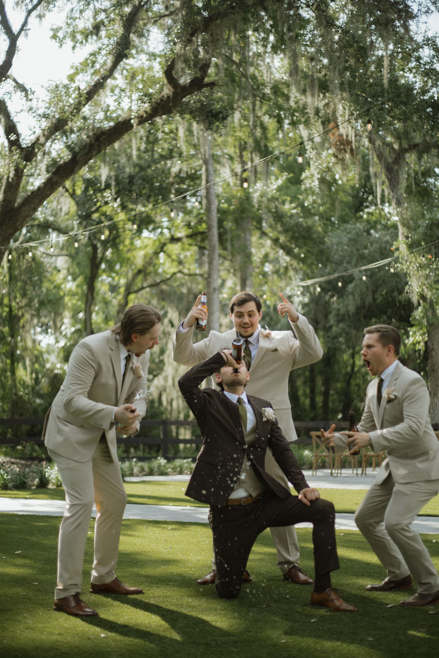 Groomsmen in tan suits cheer and hold drinks while the groom in a brown suit kneels and drinks beneath oak trees at a wedding.