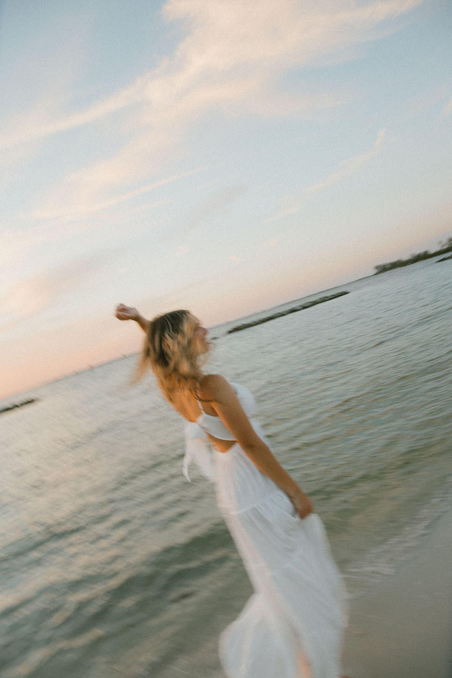 Woman in a white dress spins with an arm outstretched on the shoreline under a soft pastel sunset sky.