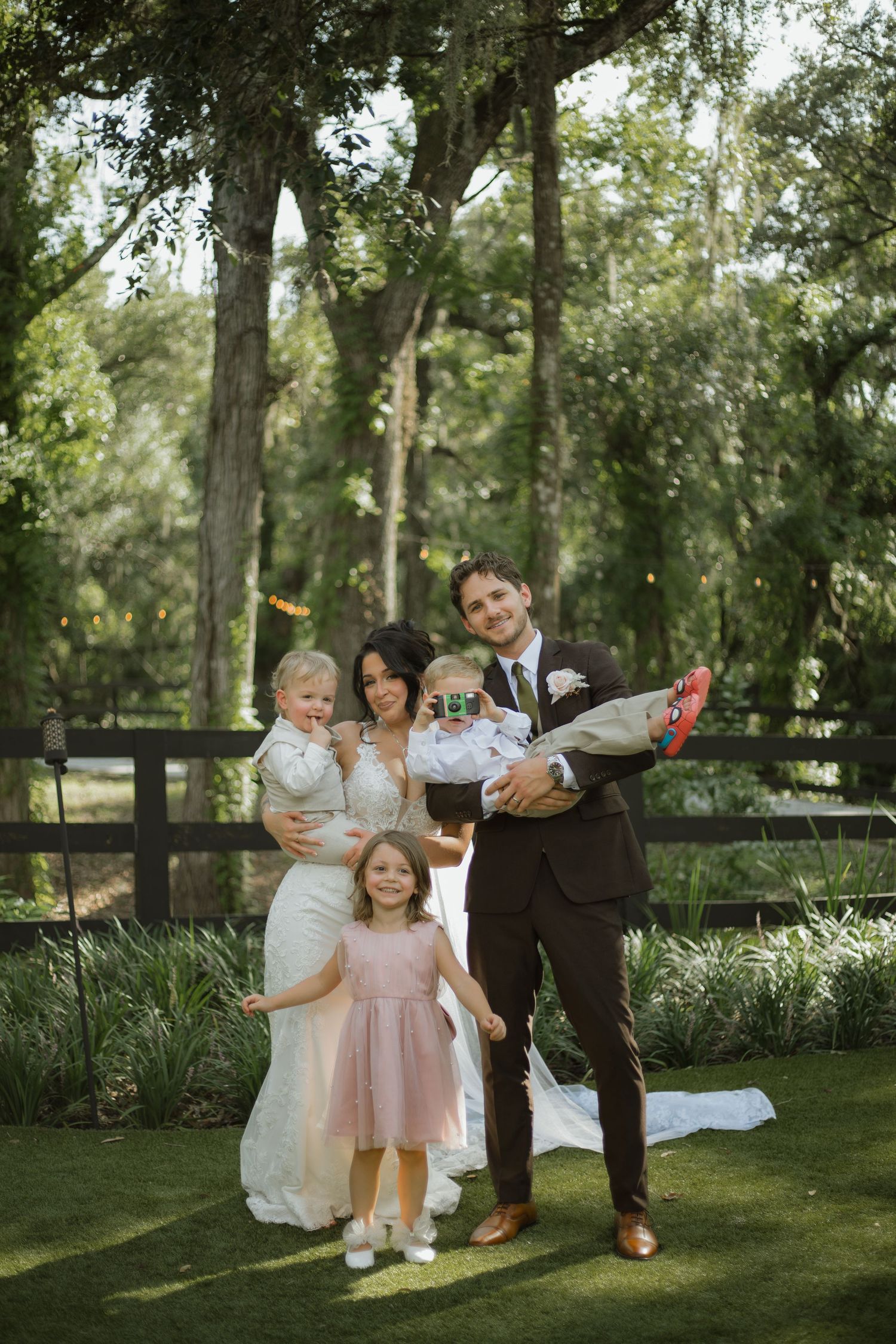 Bride and groom hold two young children on a lawn while a little girl in a pink dress reaches toward them at an outdoor wedding.