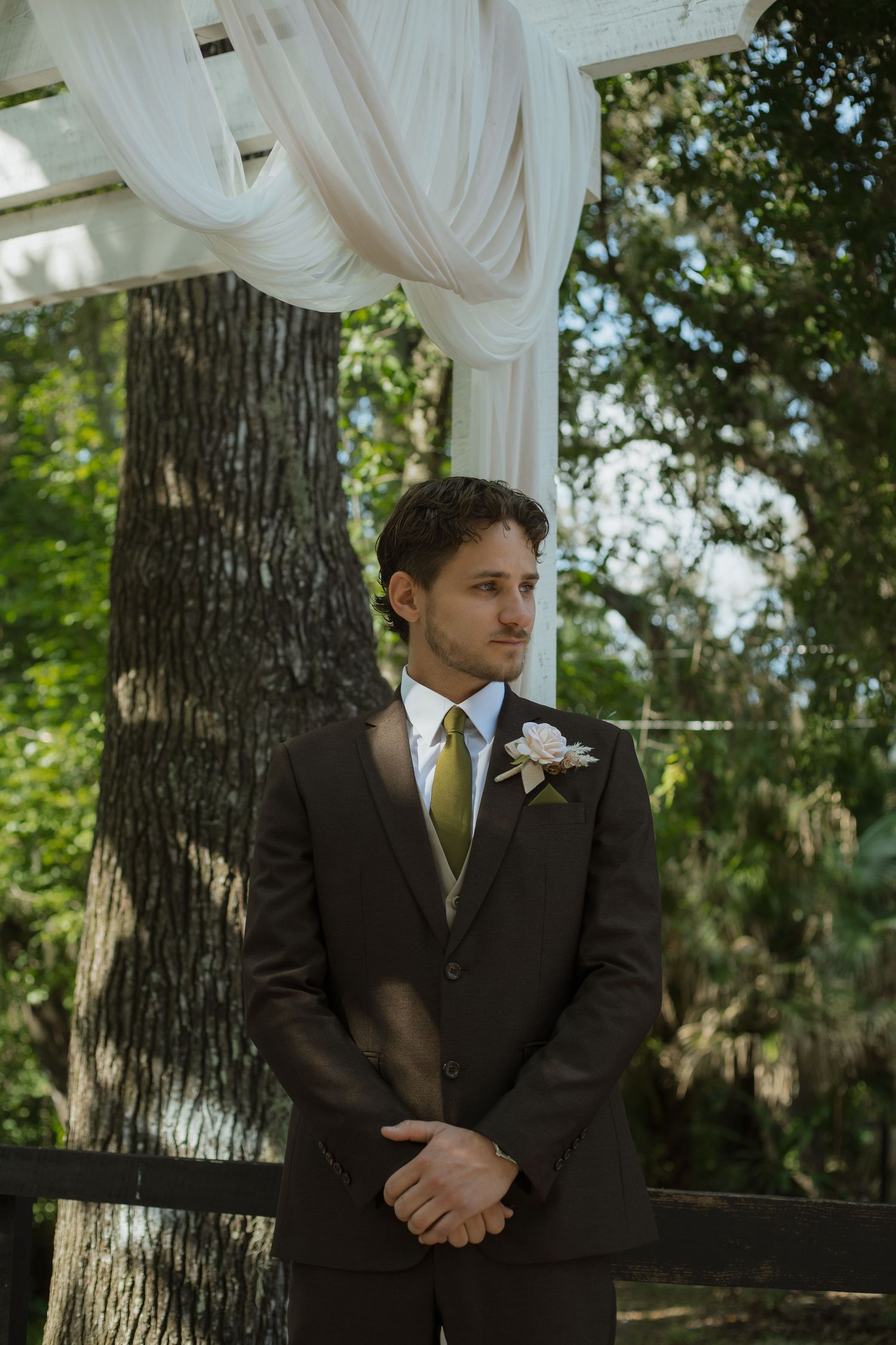 Groom in a brown suit with olive tie and white boutonniere stands with hands clasped beneath a draped white wedding arch by a tree.