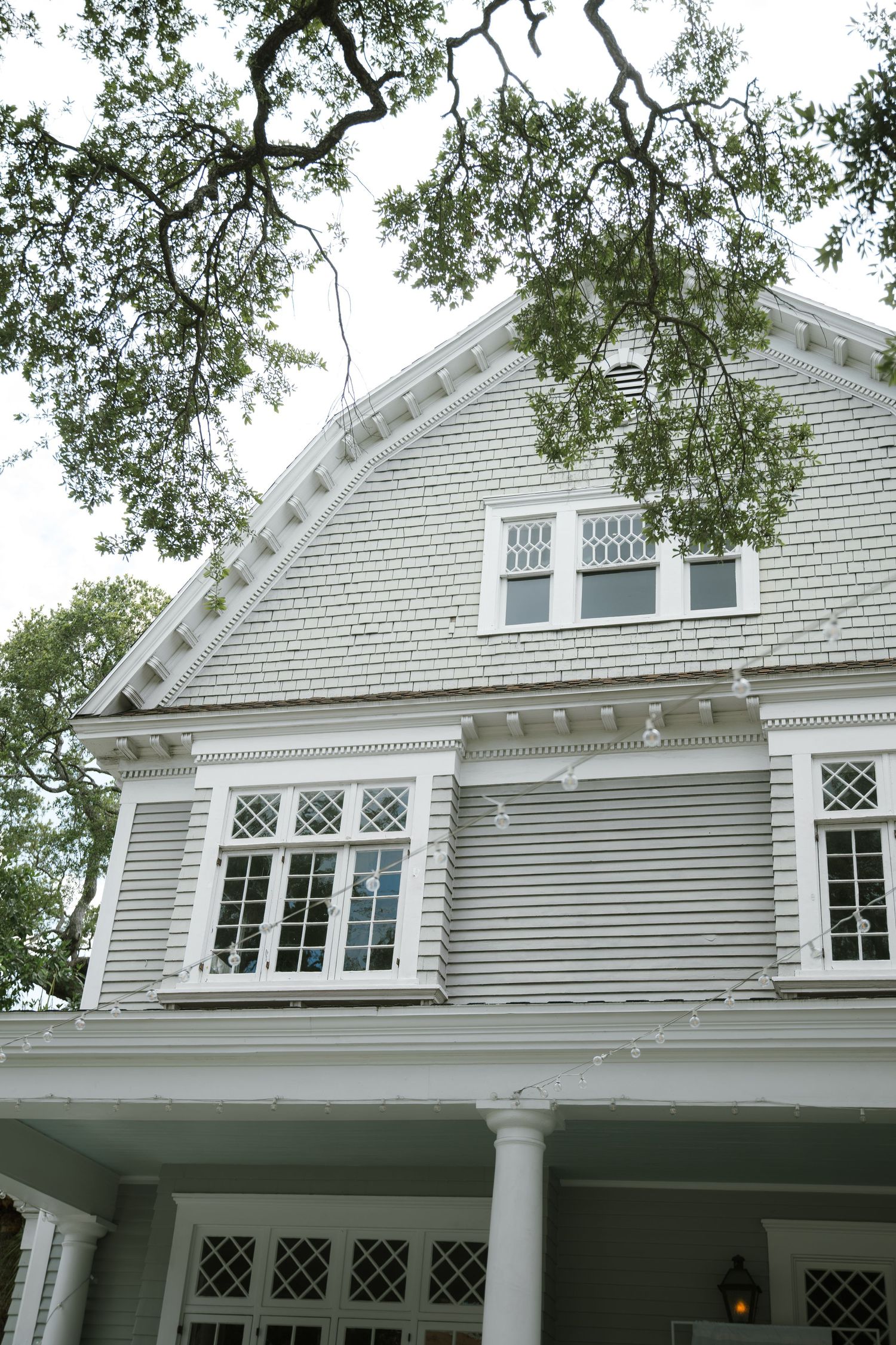 Gray shingled house with white trim, multi-pane windows and a columned porch beneath the branches of a large oak tree.