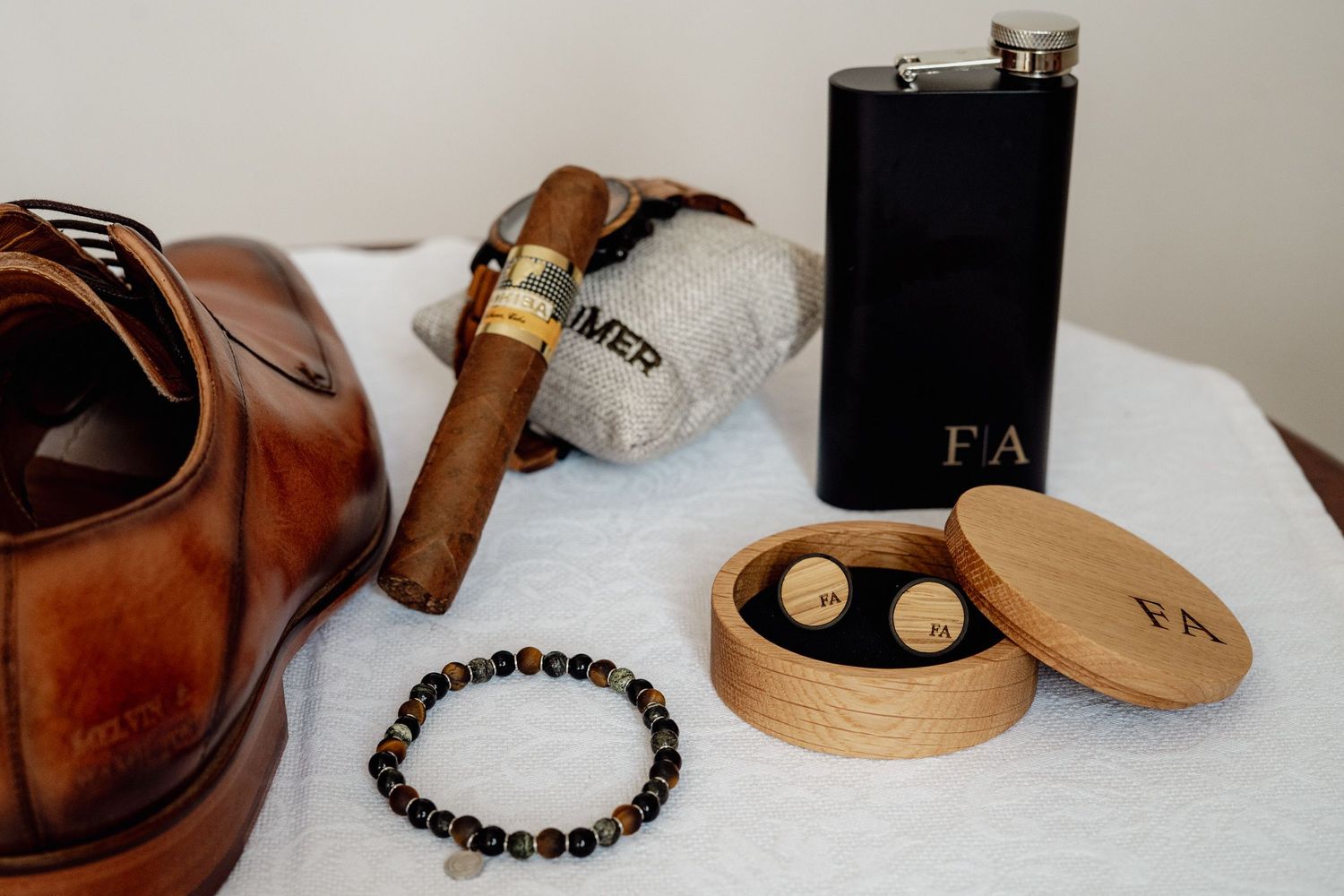 Wedding day groom accessories flat lay with leather shoes, cigar, flask, cufflinks, and bracelet.