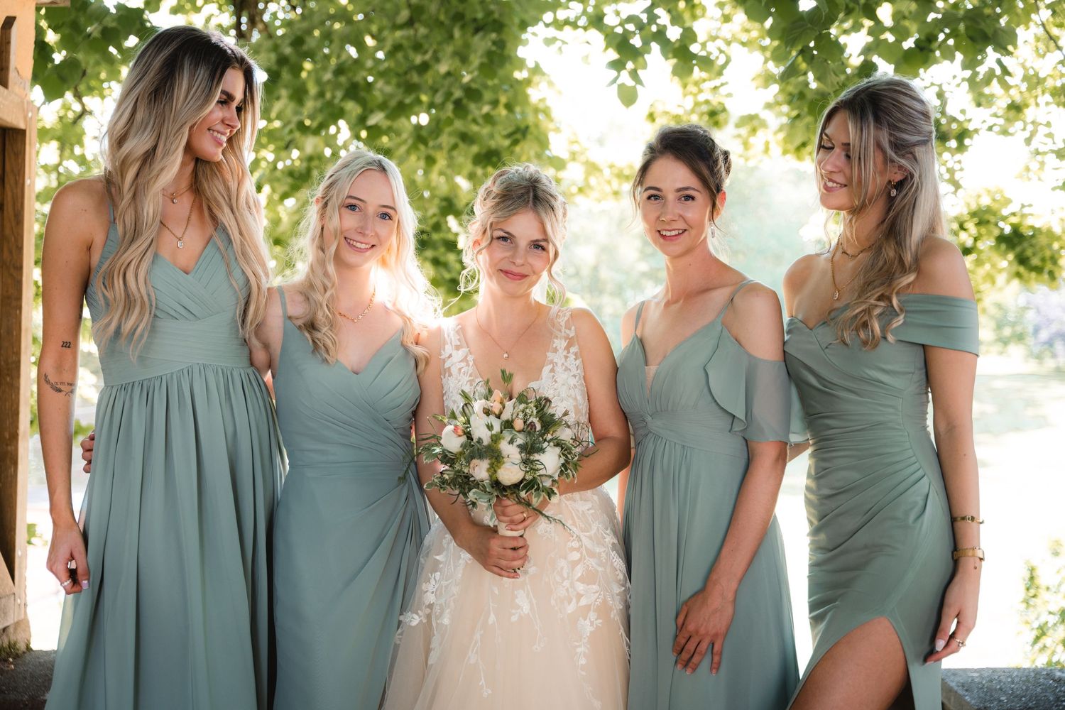 Bride in white lace gown poses with four bridesmaids in sage green dresses outdoors.