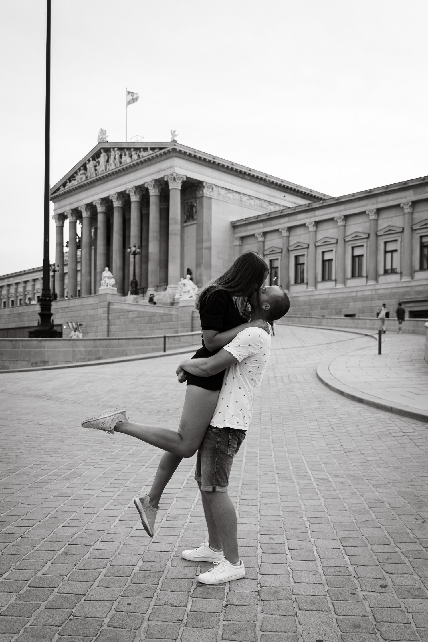 Black and white photo of couple embracing in front of Vienna Parliament building.