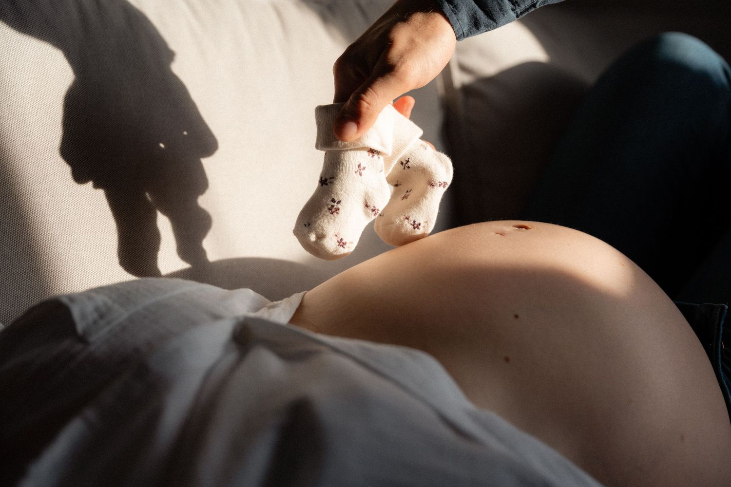 Pregnant woman holding a small stuffed animal toy above her bare belly in warm sunlight.