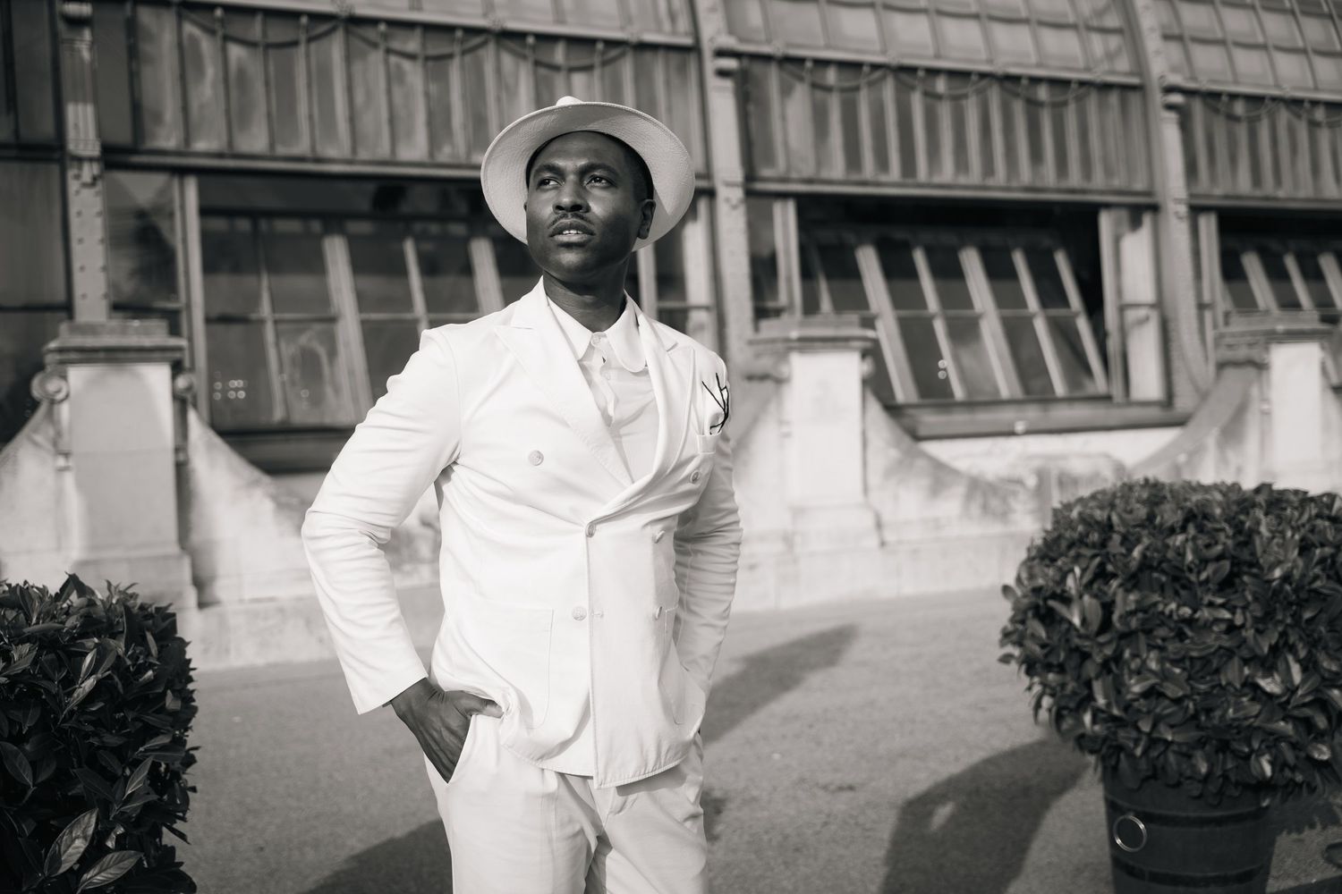 Stylish man in white suit and hat posing outdoors in black and white photo.