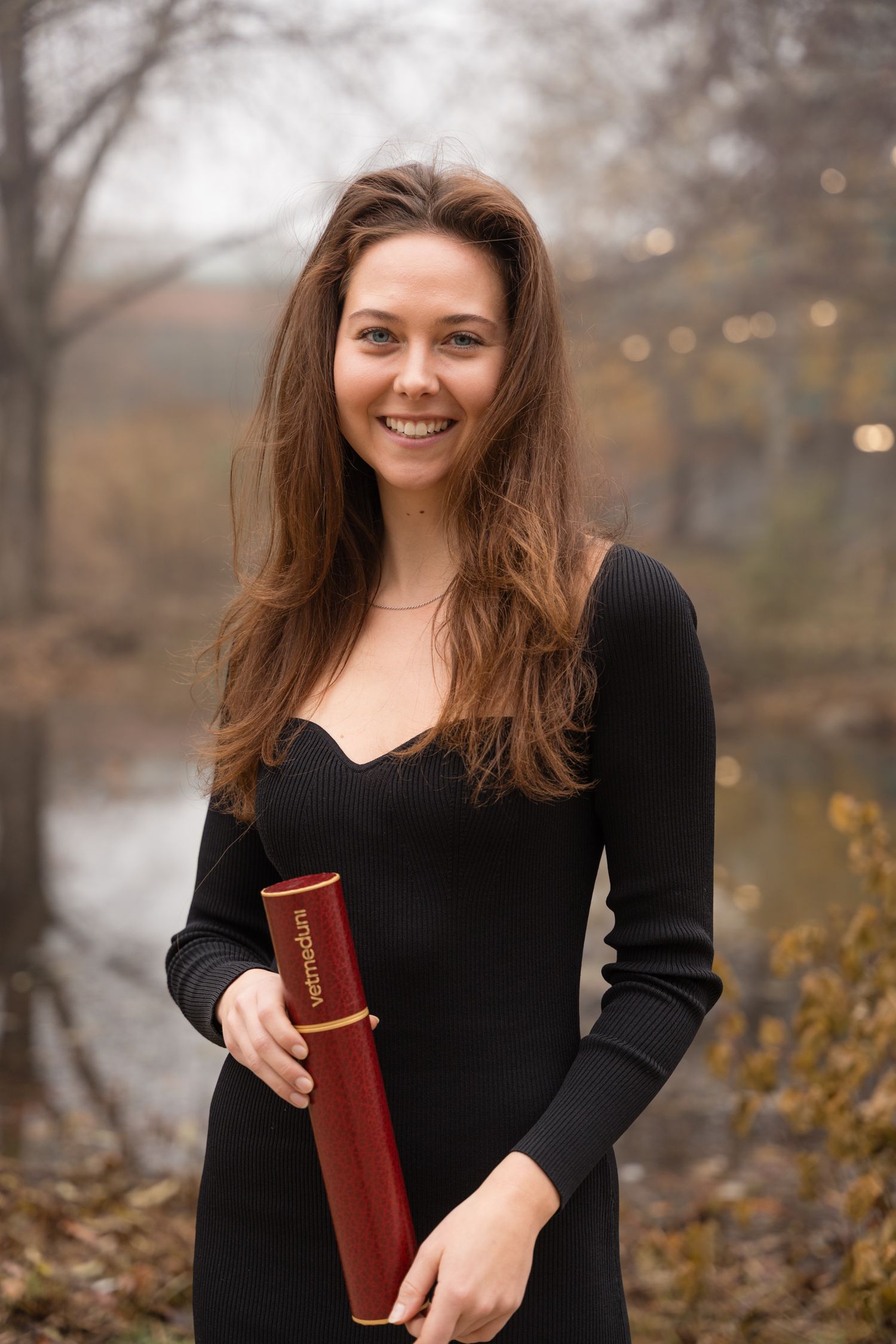 Smiling young woman with long brown hair holding a red diploma outdoors in autumn setting.