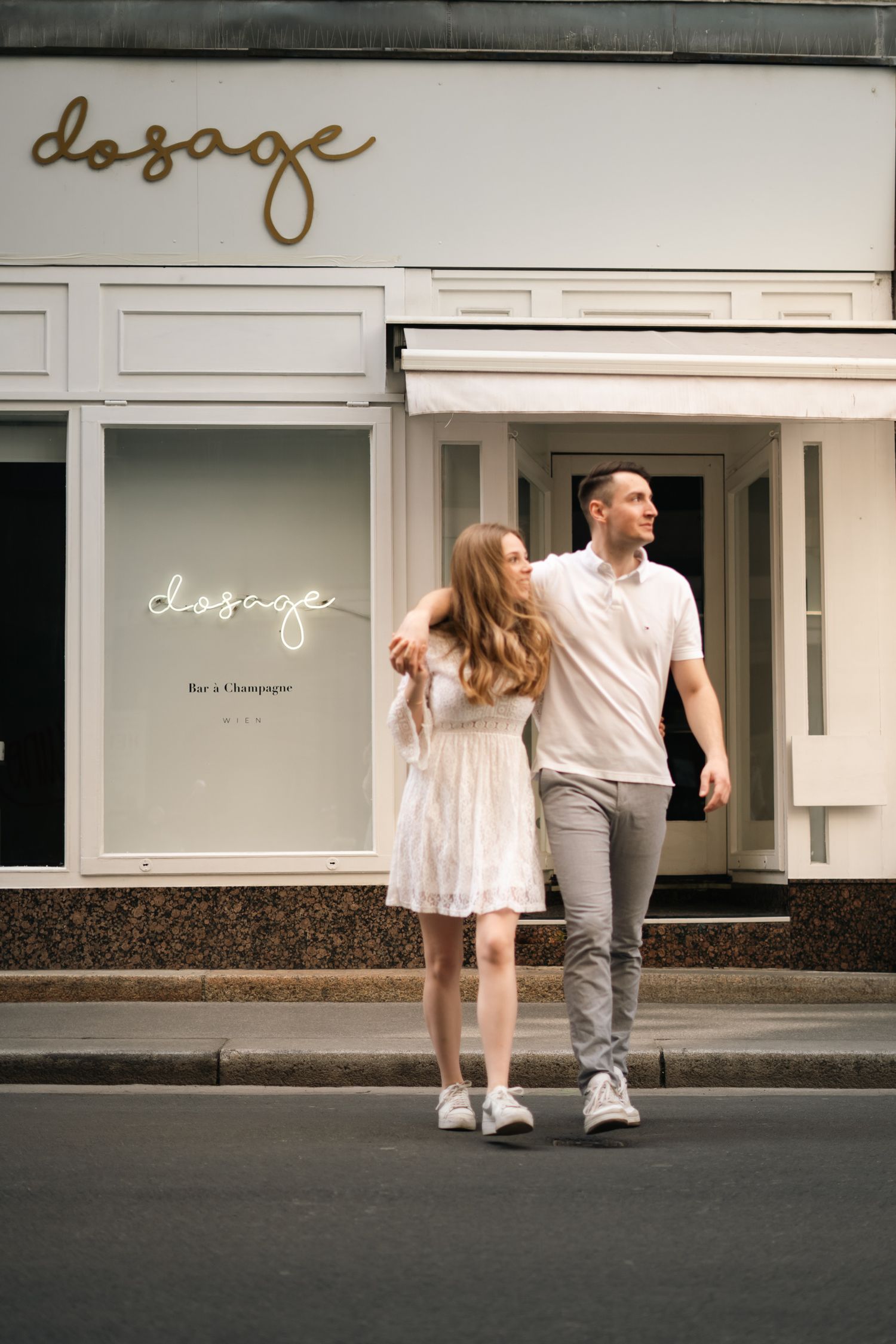 Couple walking arm in arm outside a champagne bar called Dosage on a city street.