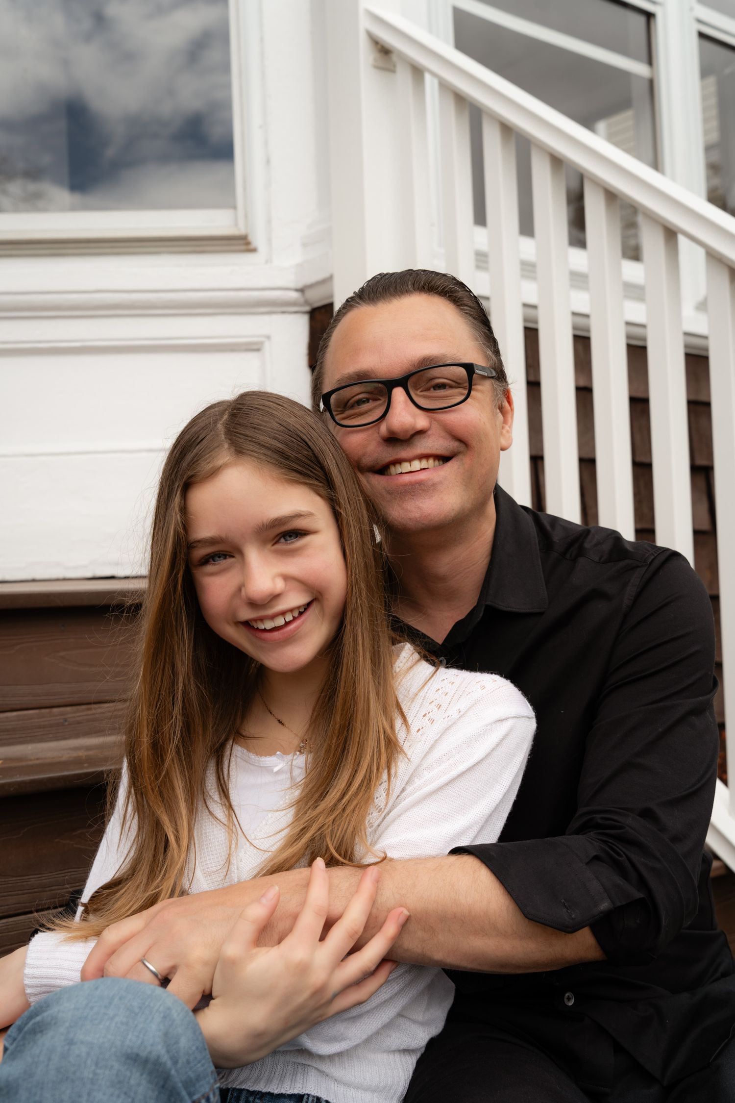 A smiling man with glasses hugs a young girl on porch steps.