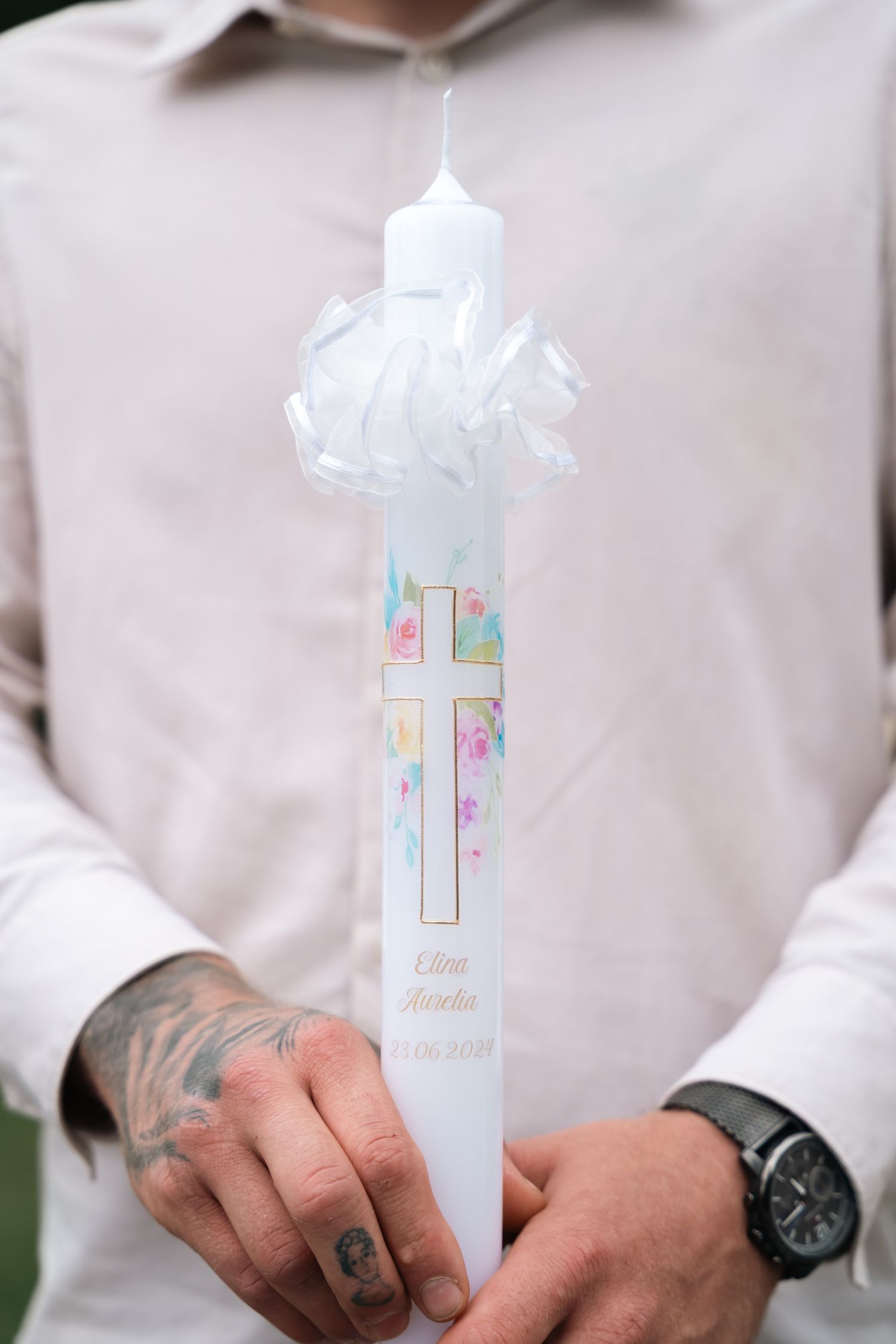 Tattooed man holding a decorative baptism candle with a cross and floral design.
