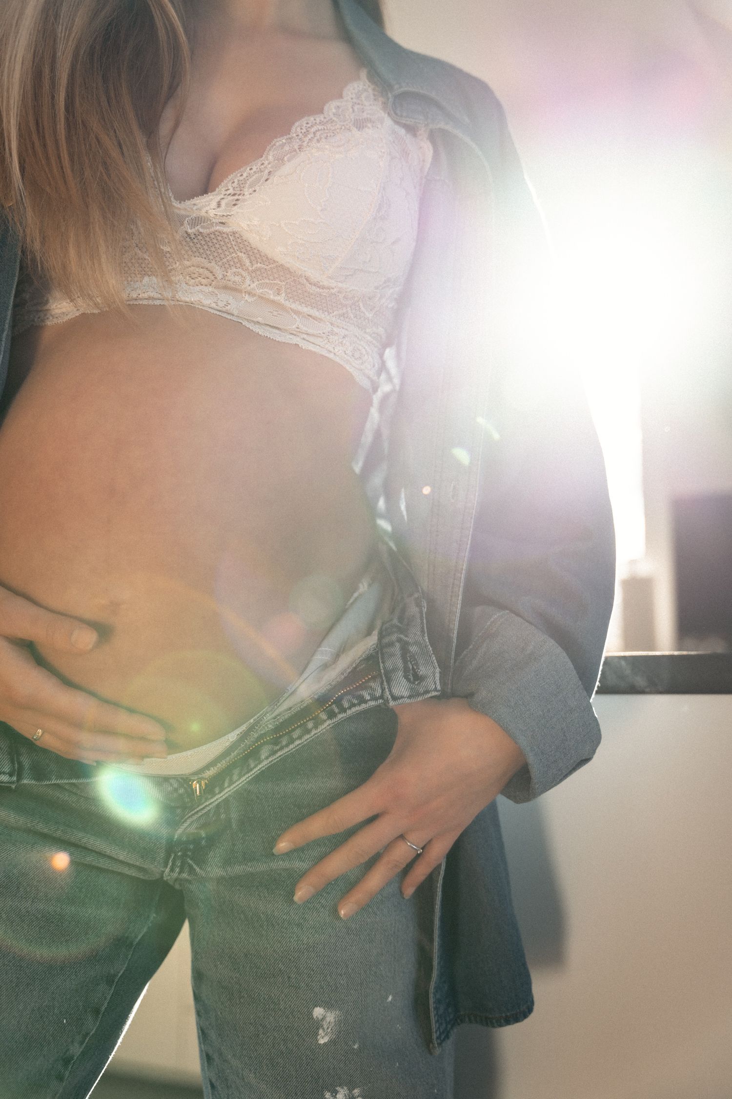 Close-up of pregnant woman in white lace bra and denim jacket holding baby bump with lens flare.