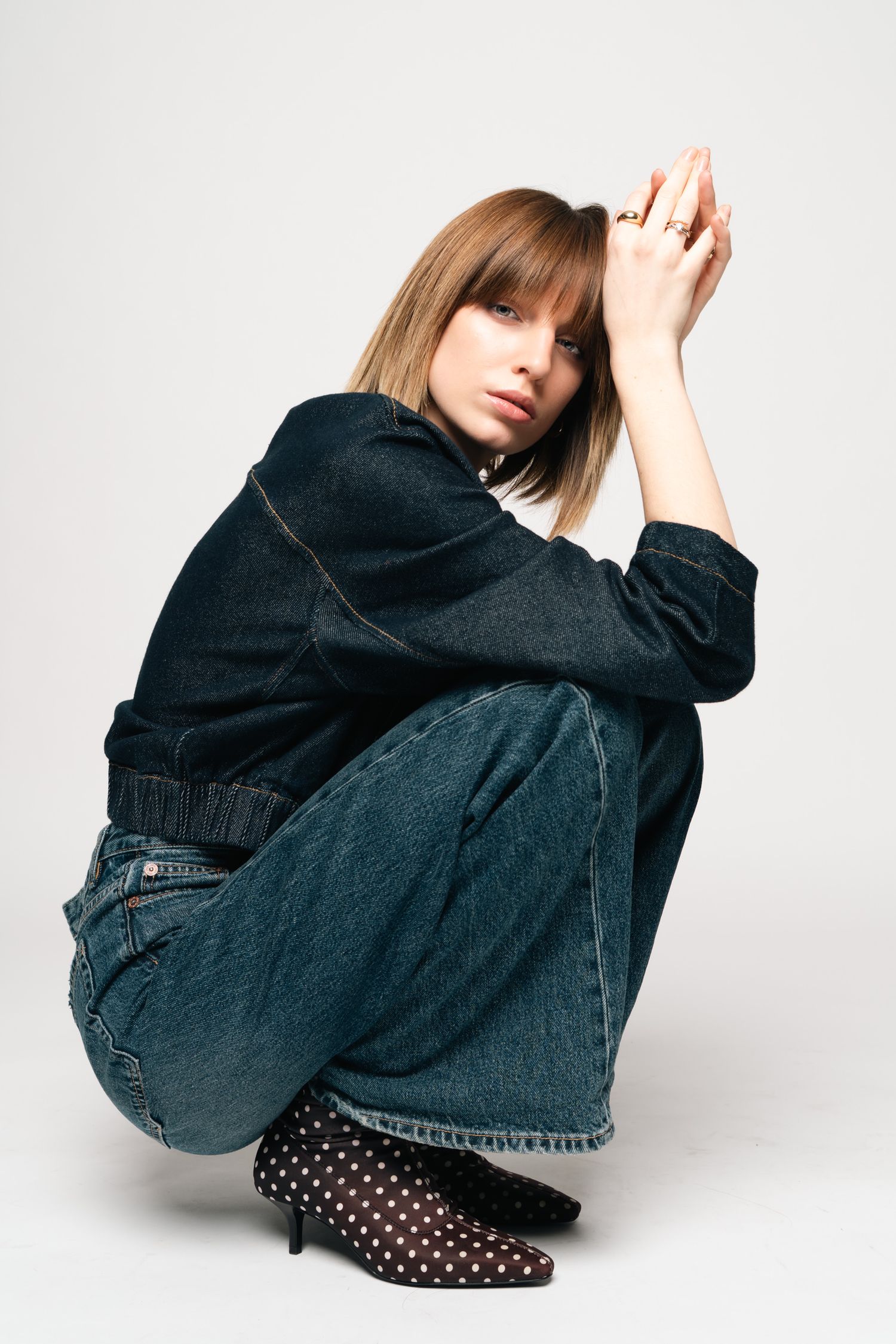 Woman with bangs sitting crouched wearing dark denim jacket, wide-leg jeans, and polka dot heels on white background.