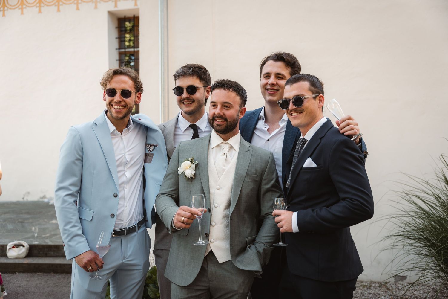 Groom in grey suit smiles with four groomsmen holding champagne glasses outdoors.
