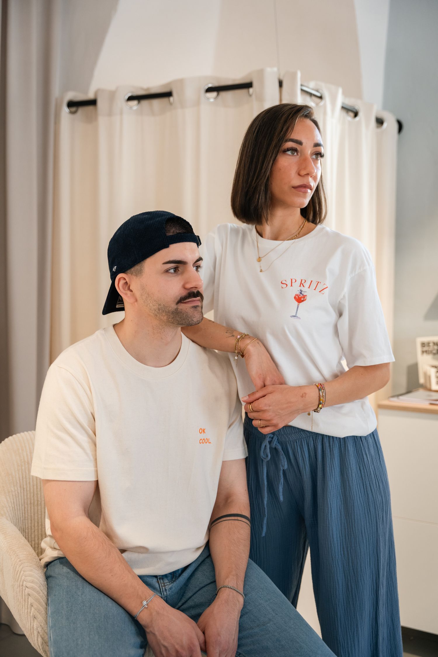 A man and woman wearing matching white Spritz graphic t-shirts pose together in a bright minimalist interior.